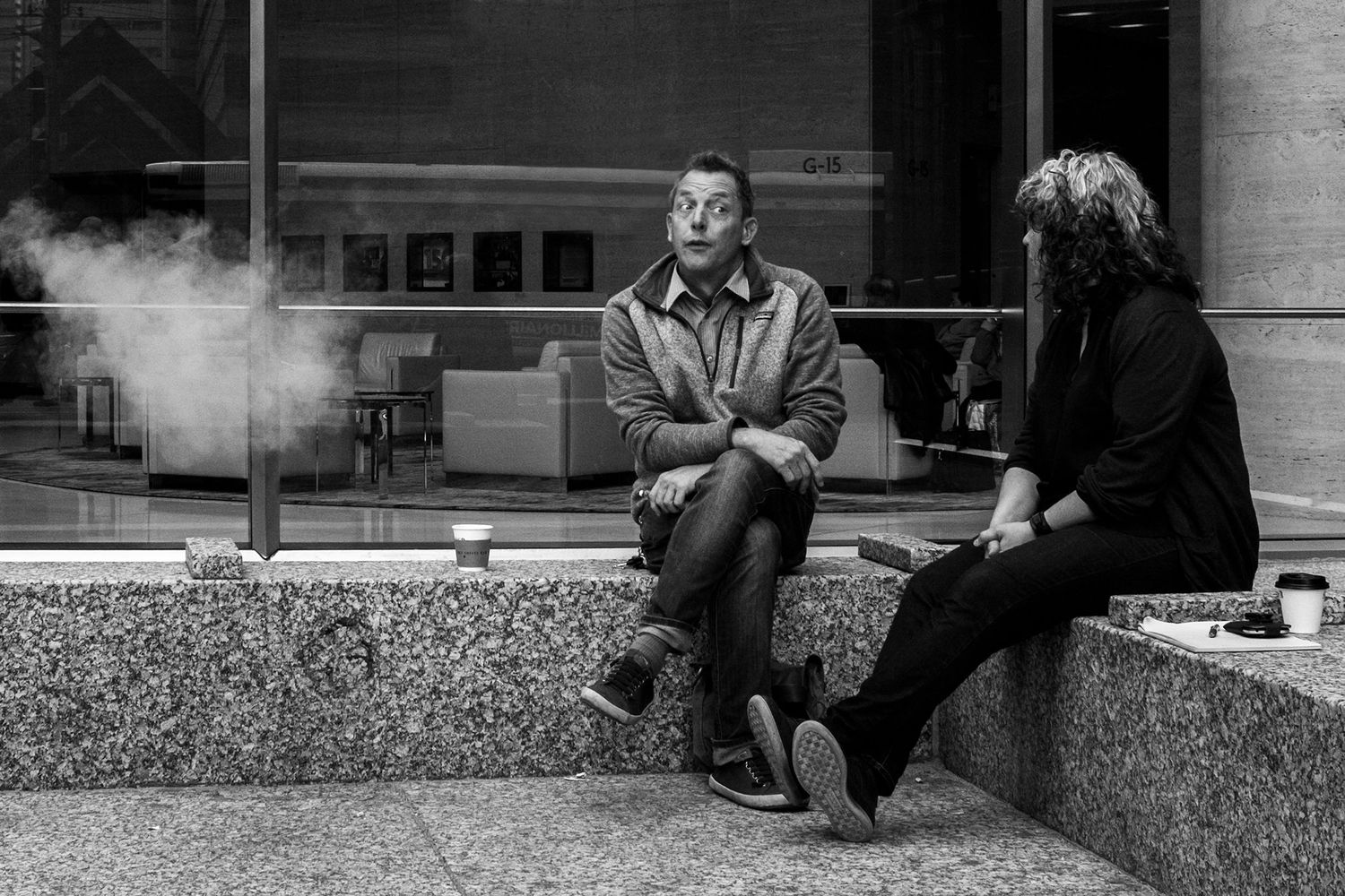 Black and white photo of two people sitting on steps outside a building, one exhaling smoke.