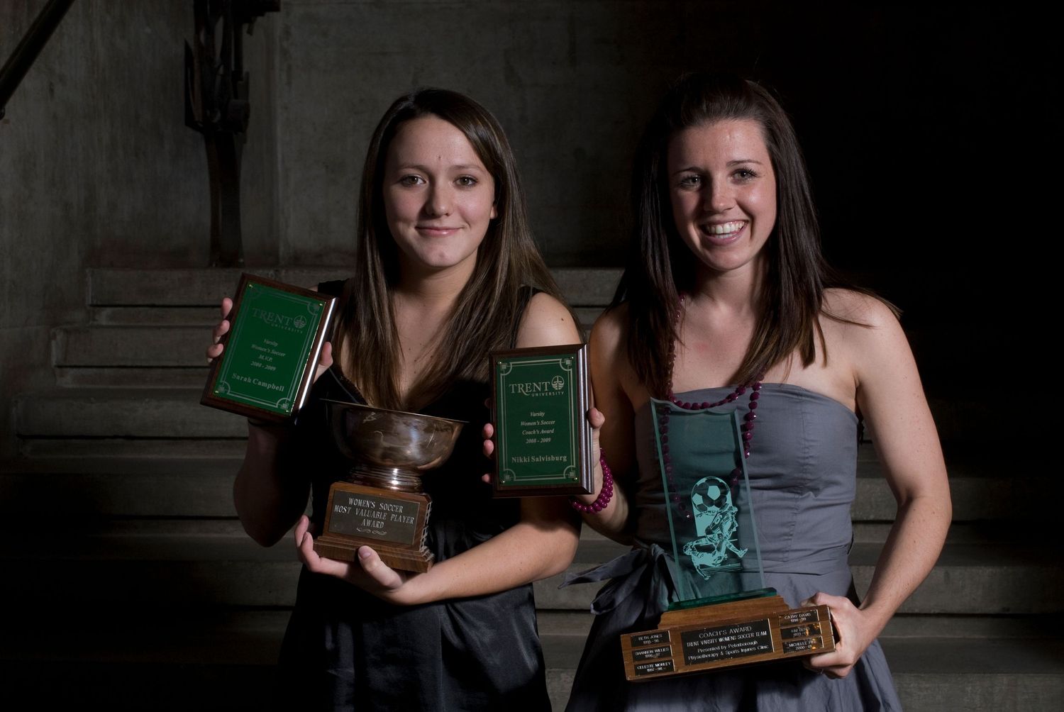 Two young women smiling and holding green awards trophies at a formal recognition ceremony.