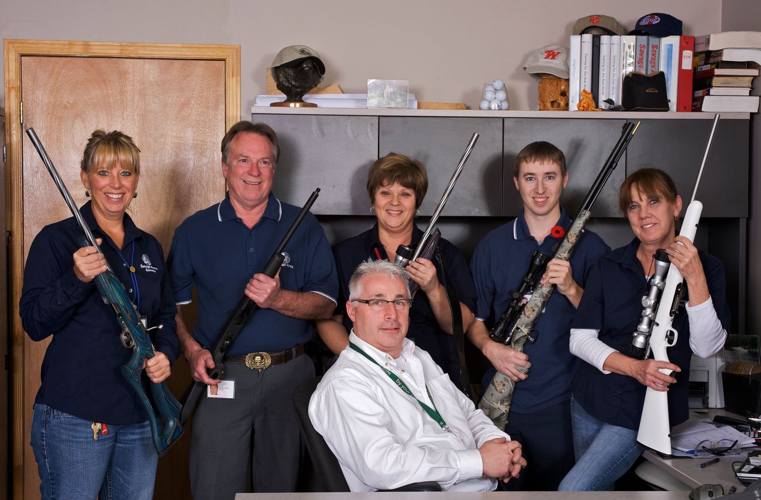 Group of six people in blue polo shirts posing with rifles in an office setting, smiling for a team photo.