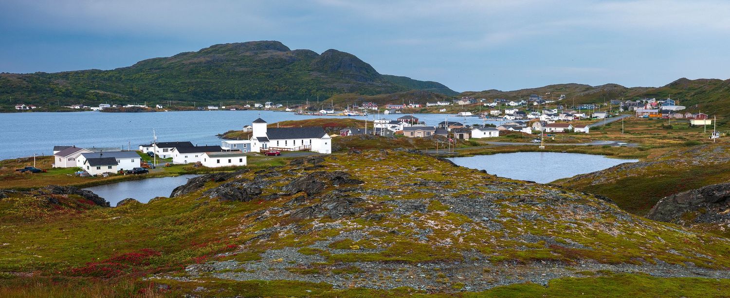 Panoramic view of a small coastal village with white buildings, rocky terrain, and mountains reflected in calm water.
