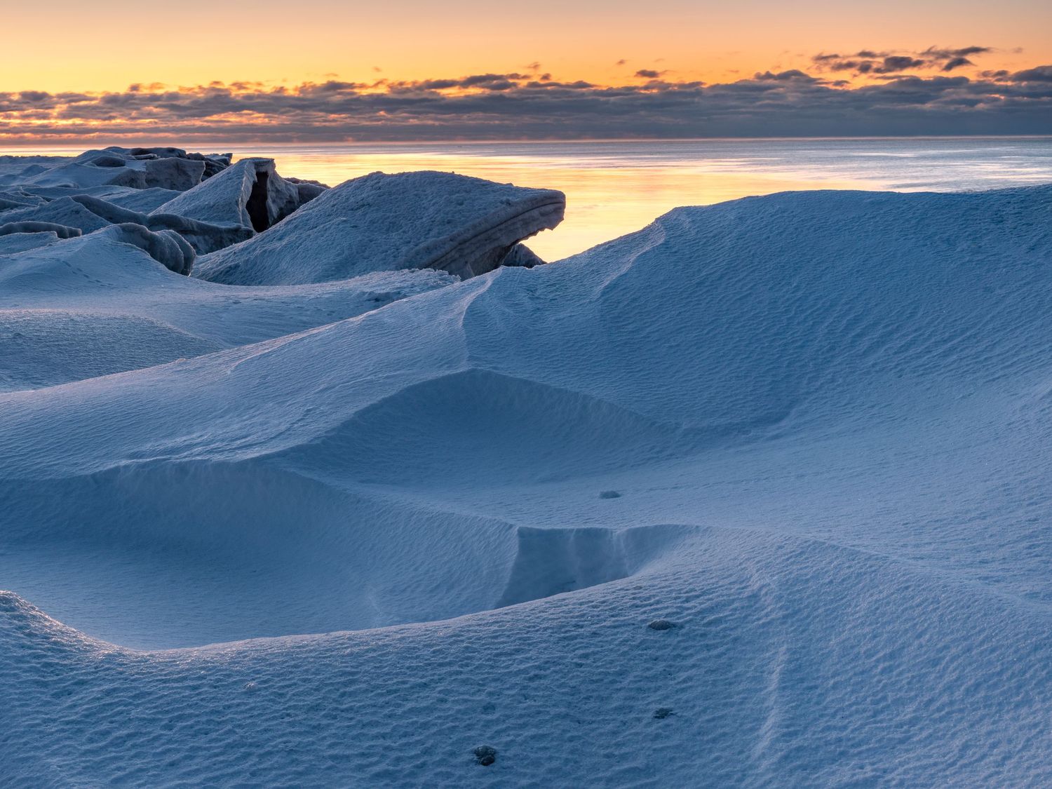 Snow-covered rocks glow in warm sunset light beside a calm Arctic sea at dusk.