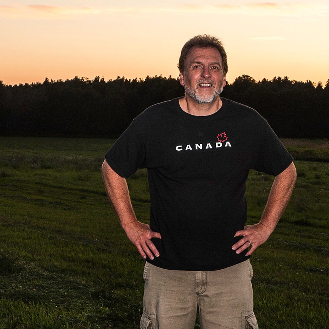 Man in black Canada t-shirt stands in green field at sunset with forest silhouette in background.