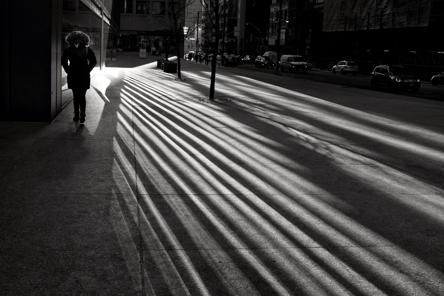 Black and white urban street photo with dramatic diagonal light rays casting long shadows on a city sidewalk with a lone pedestrian.
