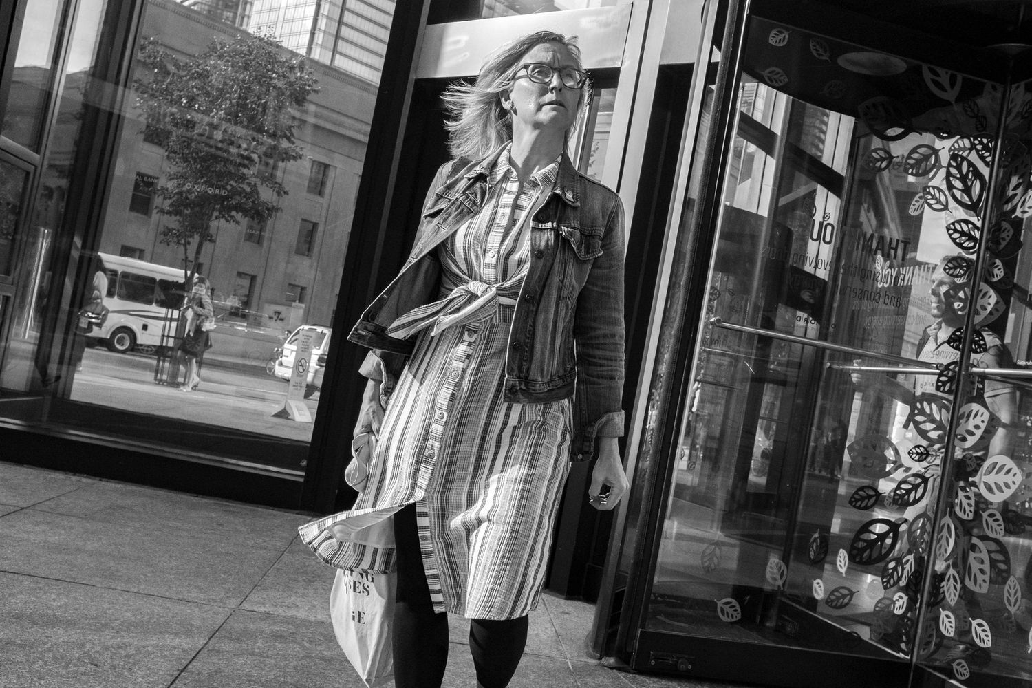 Black and white street photo of a stylish woman in striped dress and jacket walking past a glass building.