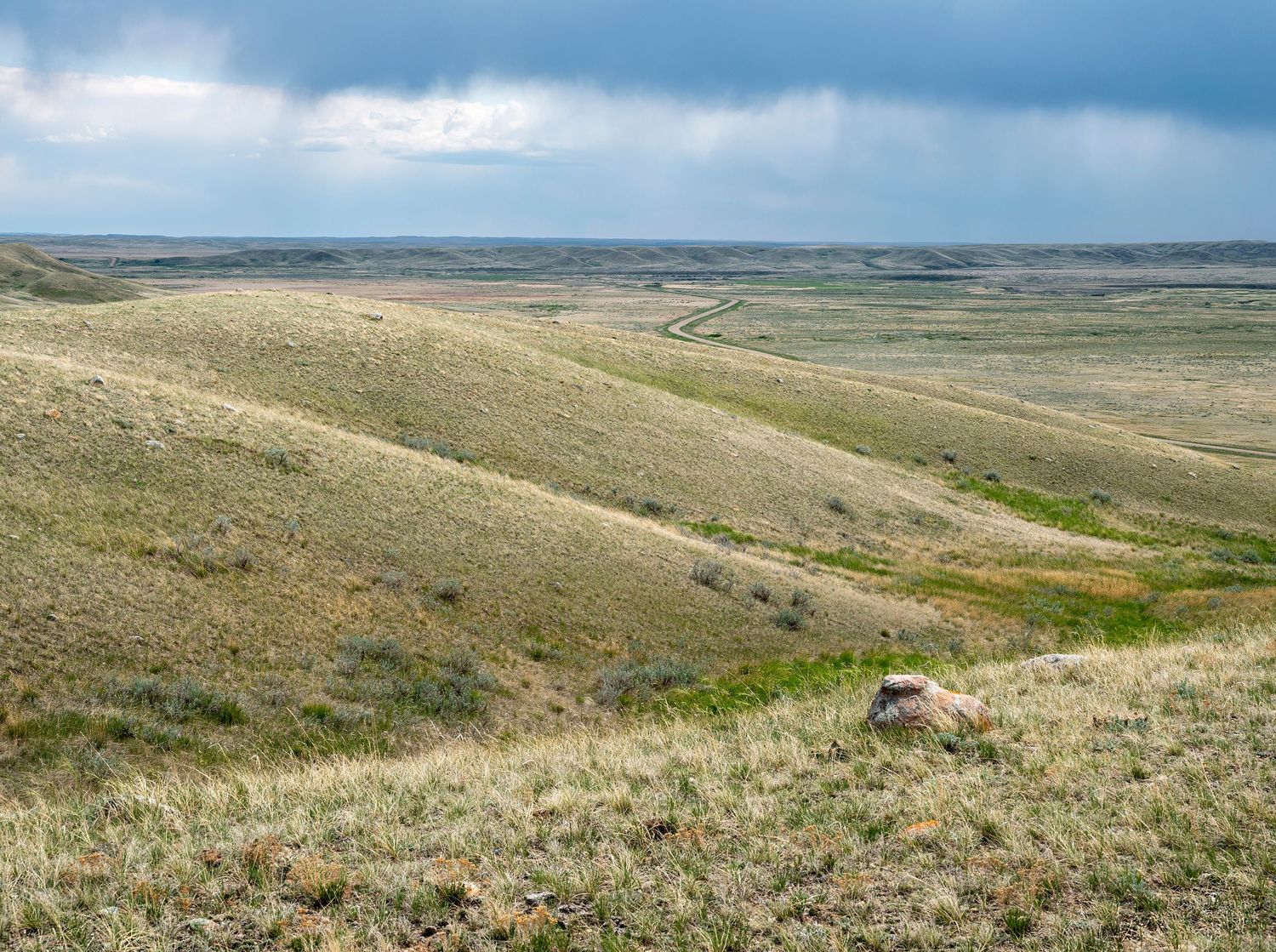 Vast rolling grassland prairie under stormy skies with gentle hills and a single rock in the foreground.