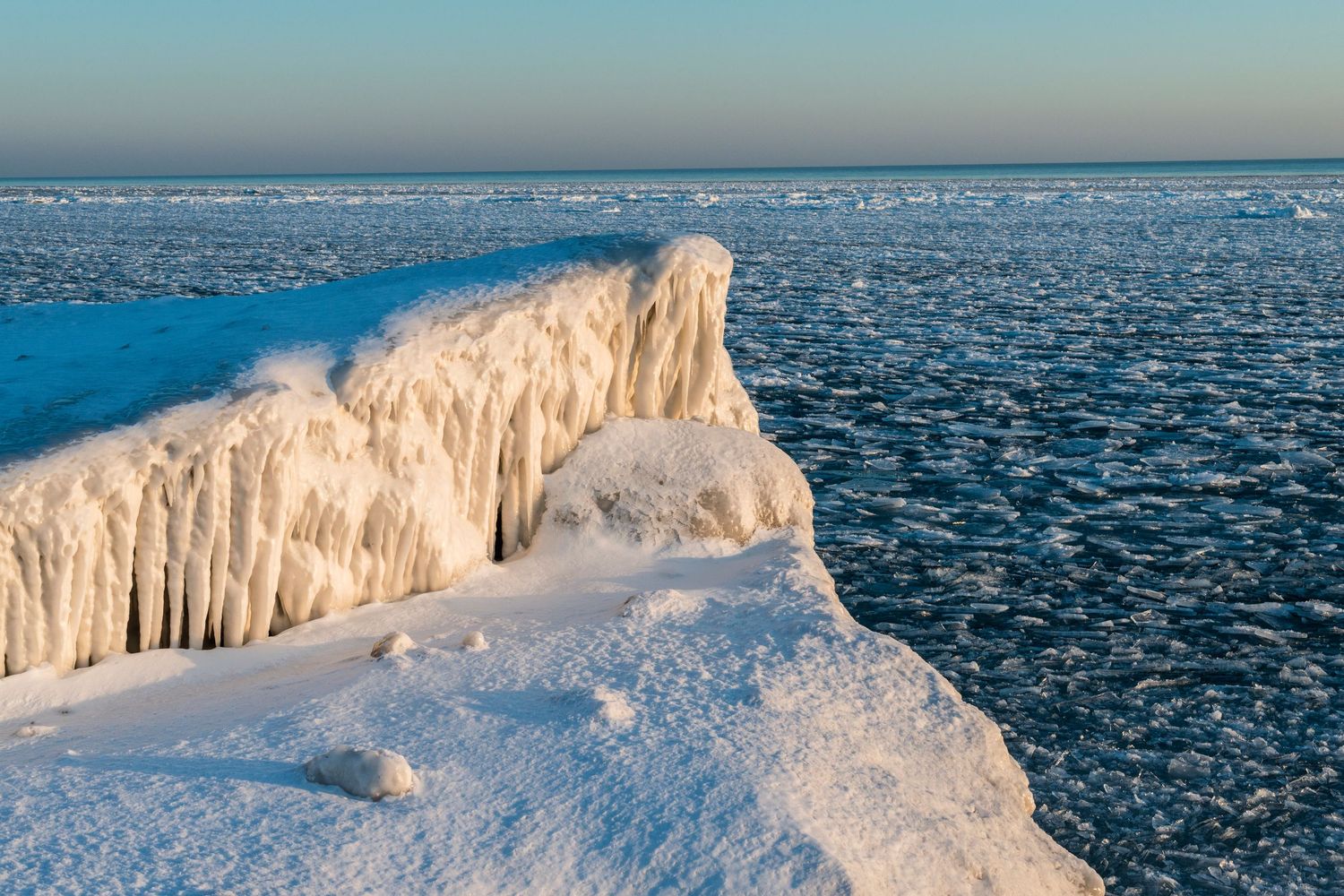 Frozen icy shoreline with dramatic ice formations and snow along a vast frozen lake under a clear winter sky.