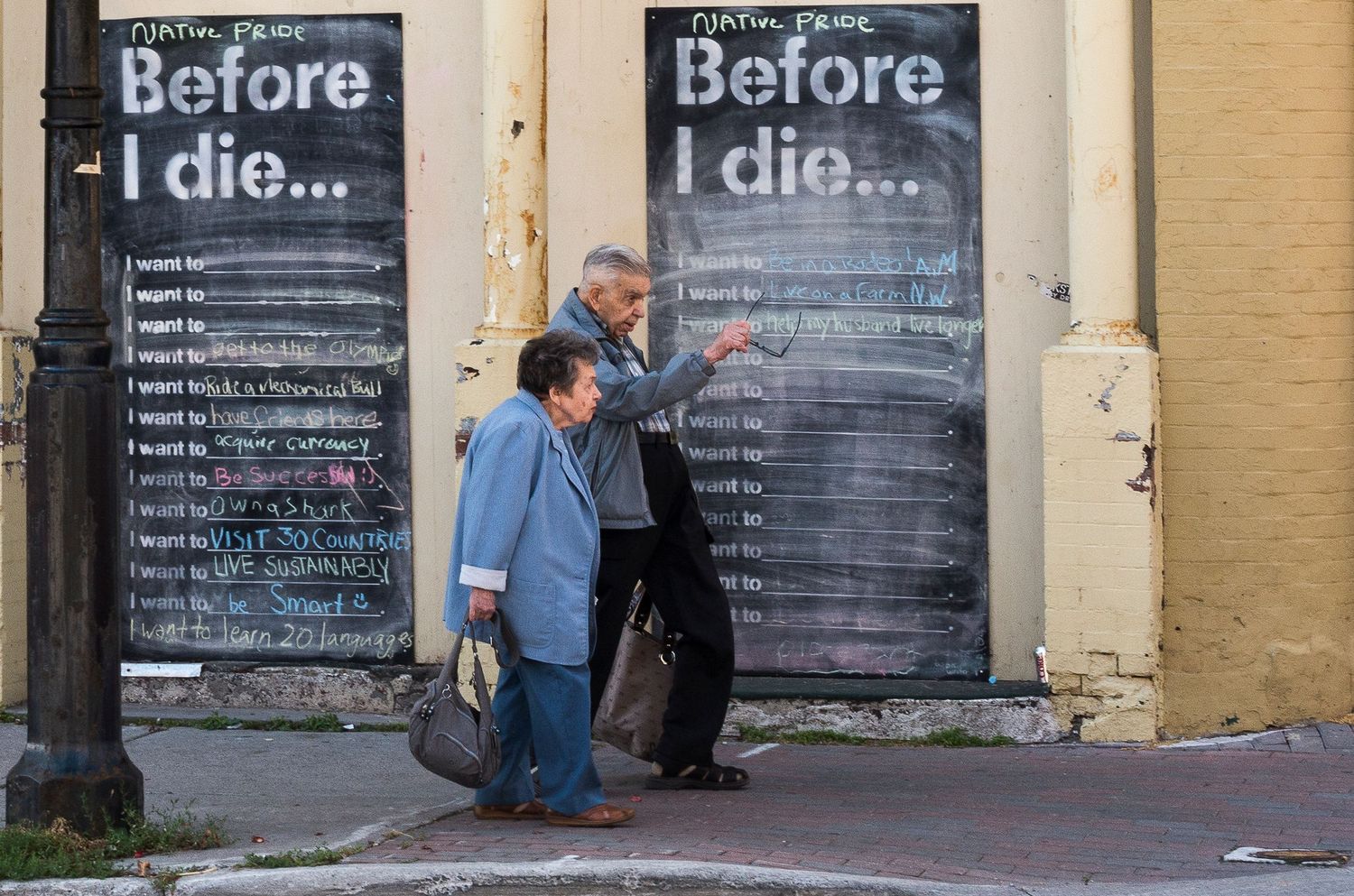 Two people walk past a large chalkboard mural reading Before I Die on a city street wall.