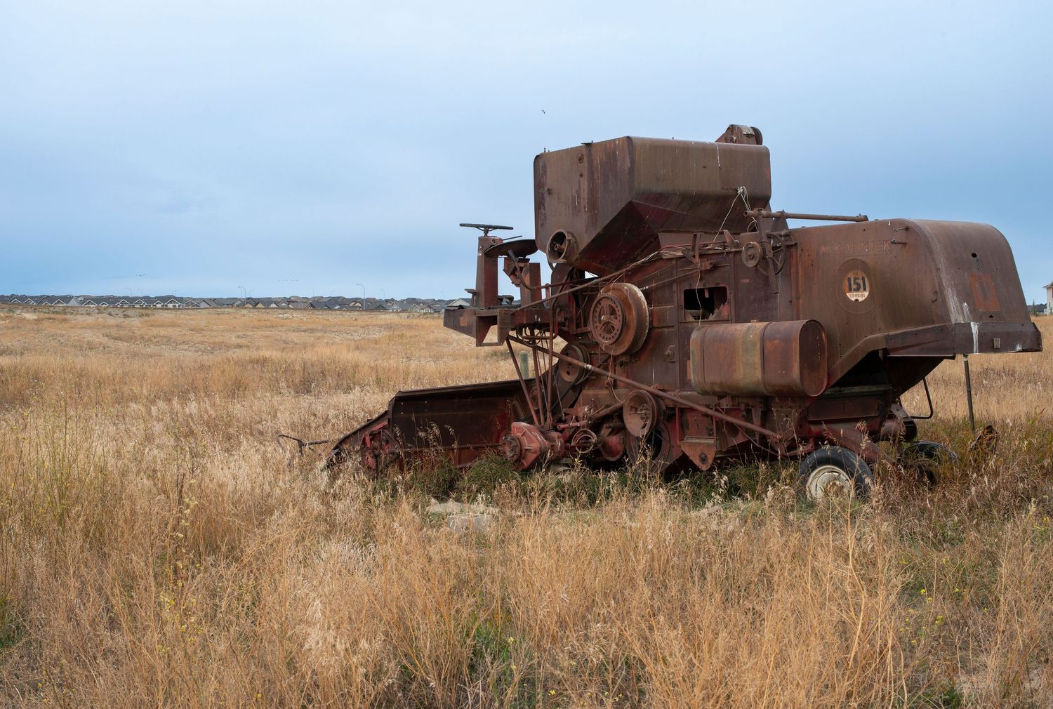 Rusty abandoned combine harvester sitting in a dry golden grass field under a cloudy sky.