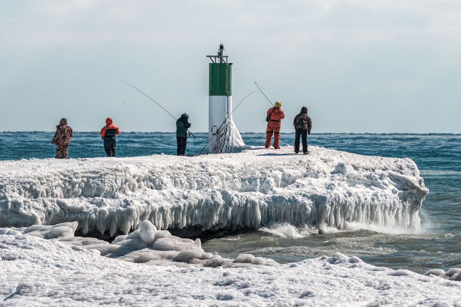 Fishermen stand on an icy pier near a green lighthouse on a frozen winter shoreline.
