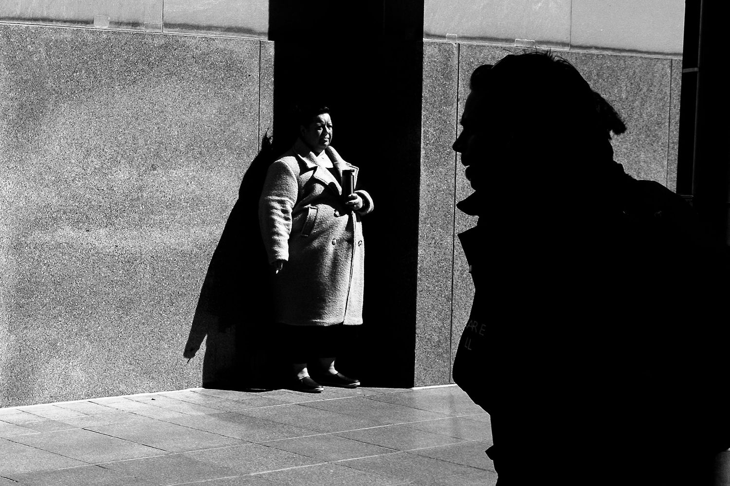 Black and white street photo of a woman in a coat standing by a wall, with a silhouetted figure in the foreground.