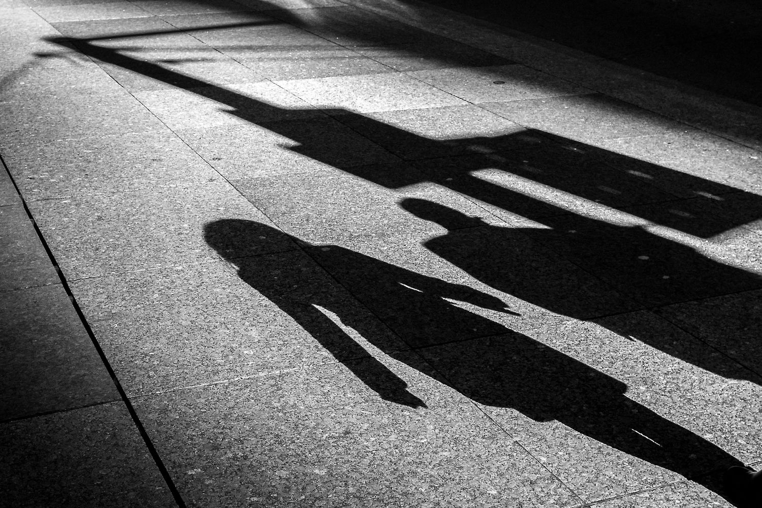 Black and white photo of dramatic shadows of a person on a sunlit sidewalk pavement.