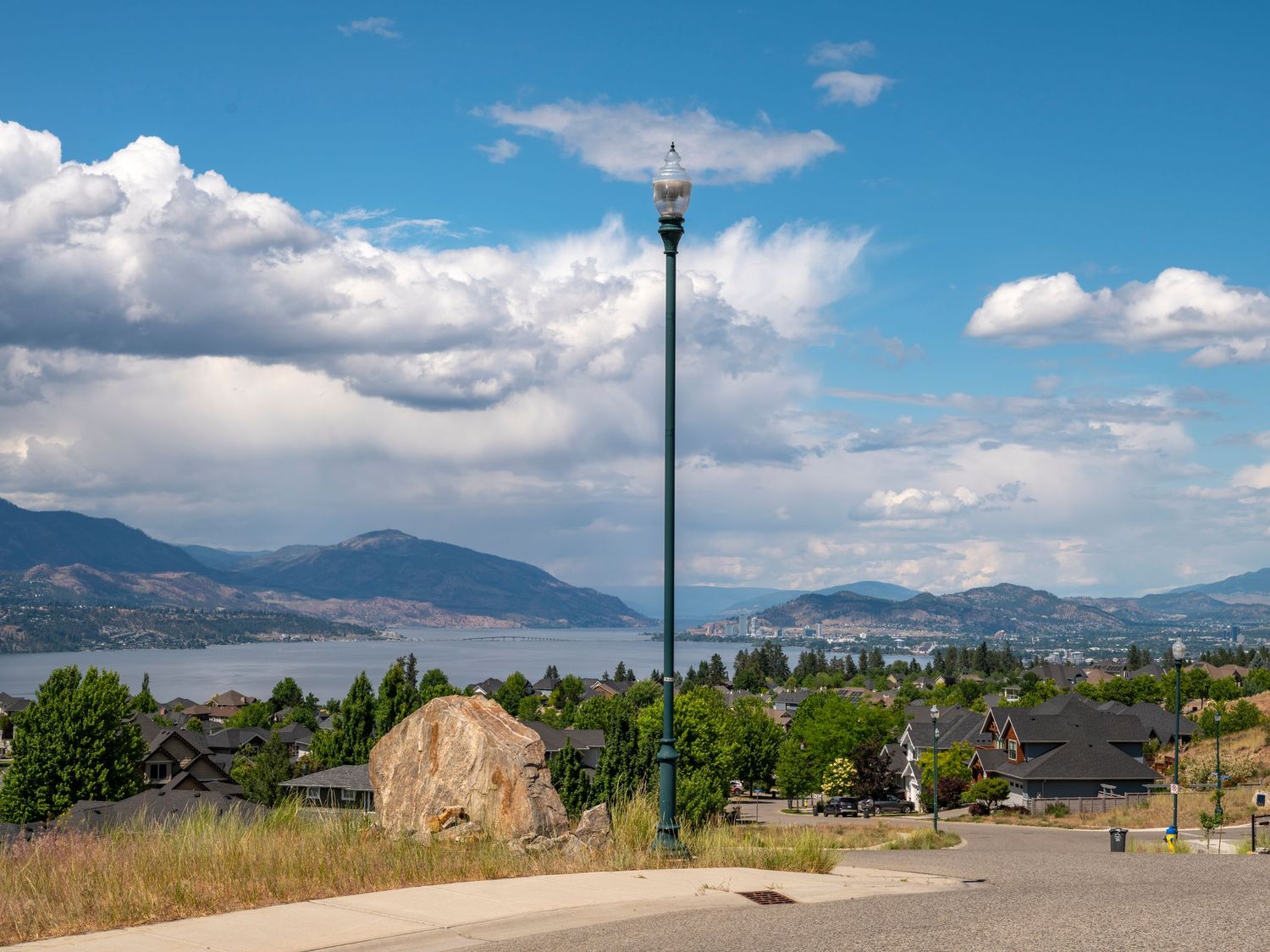 Street lamp and large boulder overlook a scenic lakeside town with mountain backdrop under blue skies.