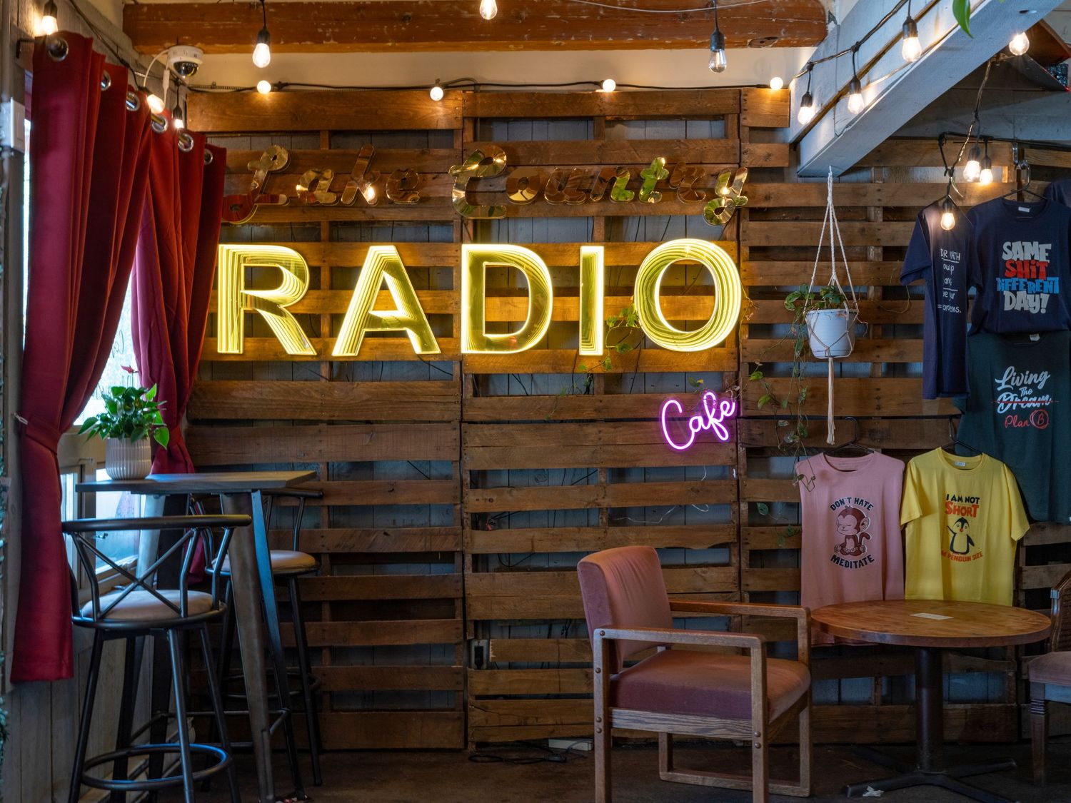 Cozy Radio Cafe interior with glowing neon sign on rustic wood pallet wall, string lights, bar stools, and colorful t-shirts.