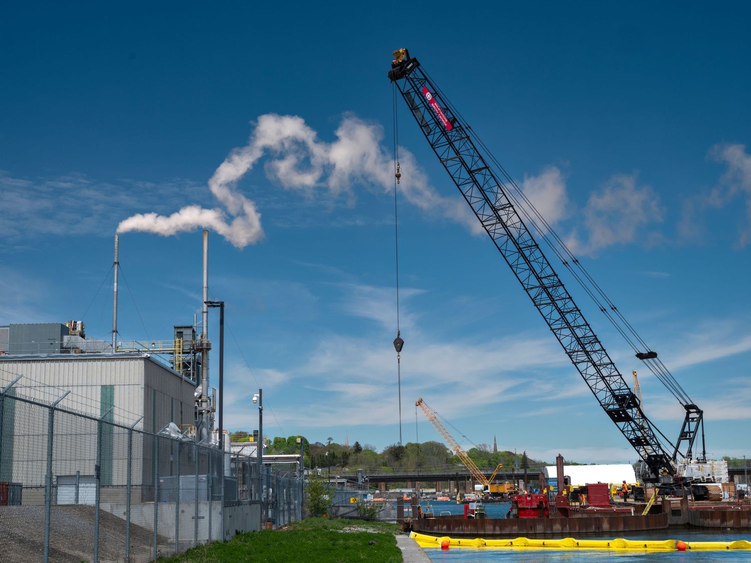 Industrial crane and factory with smoke stack against a blue sky near a waterway.