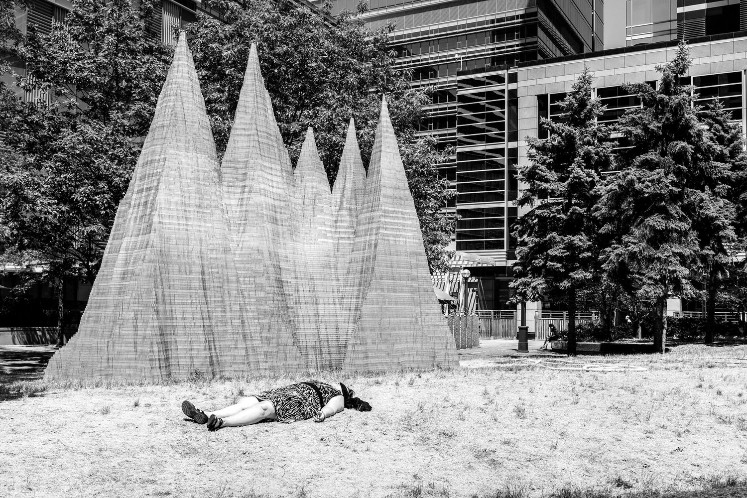 Person lying on grass near geometric pyramid wire sculptures in a black and white urban park photo.