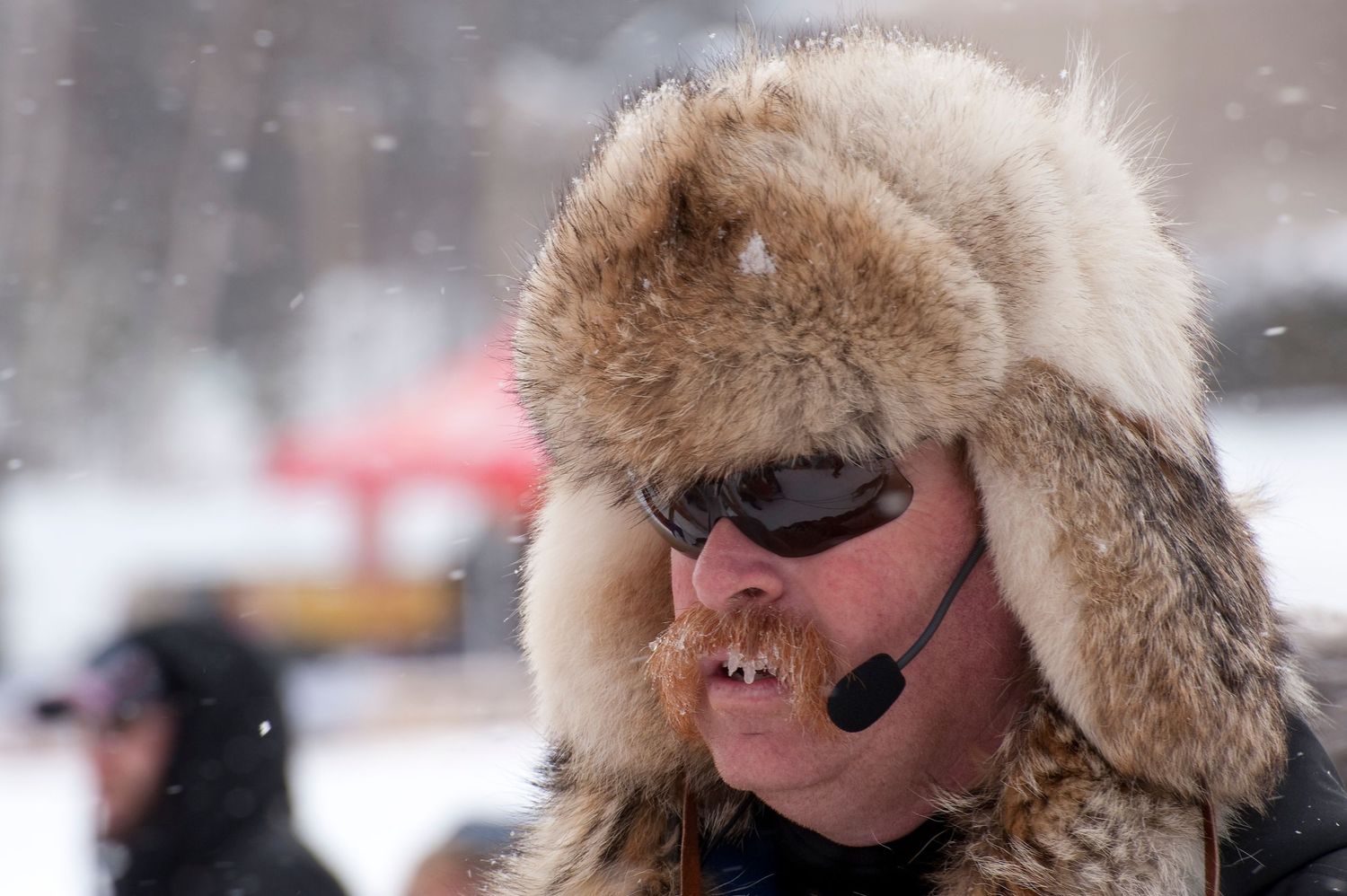 Man wearing fur trapper hat and sunglasses with headset microphone in snowy winter weather outdoors.