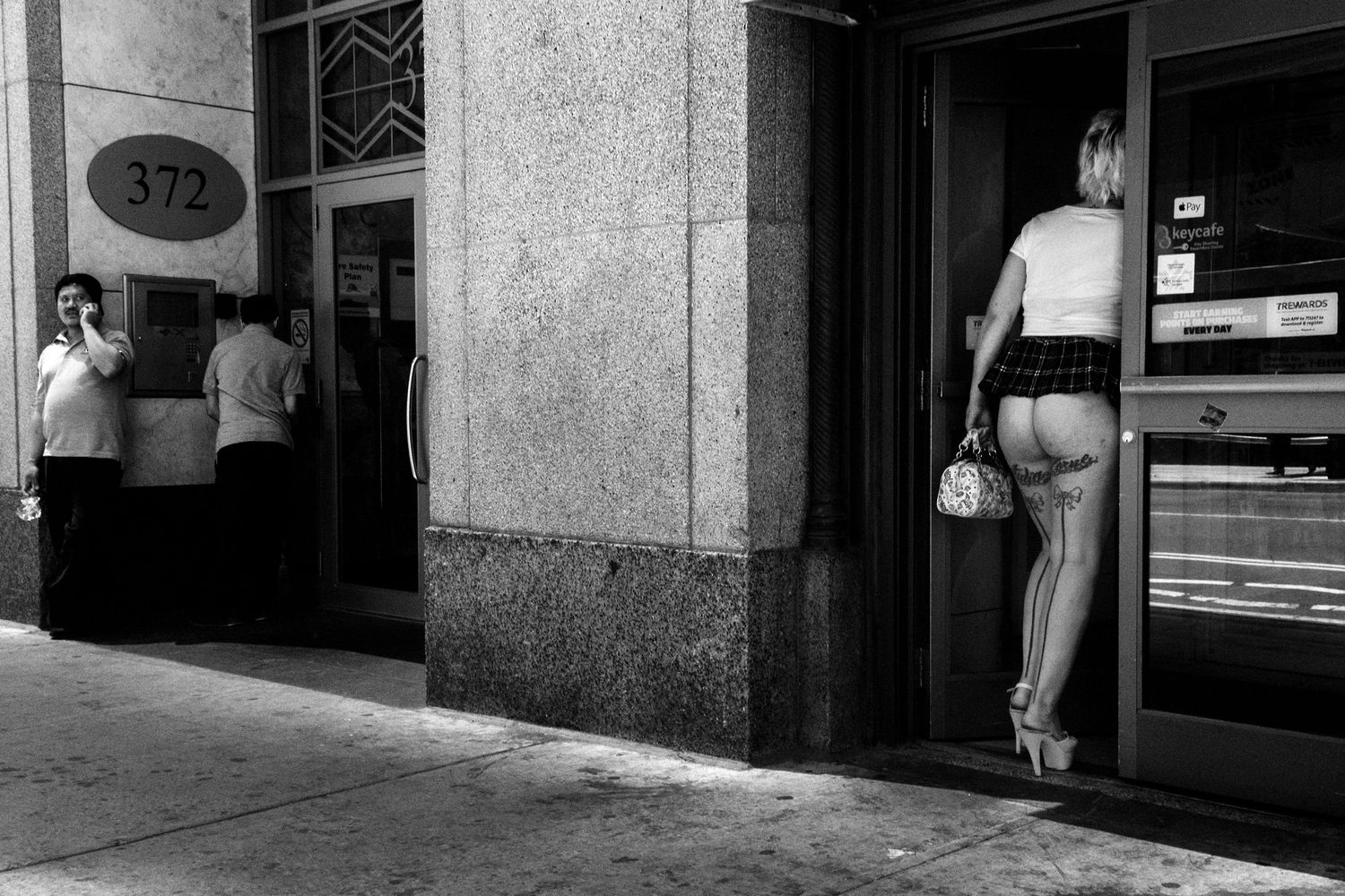 Black and white street photo of a woman entering a building at 372, with a man on phone nearby.