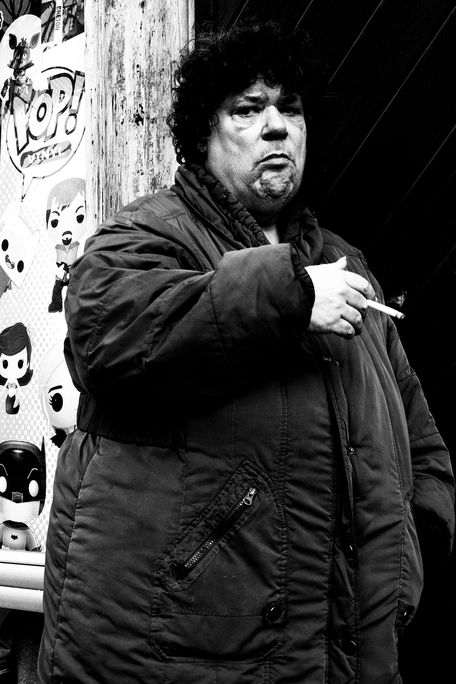 Black and white street portrait of a heavyset man smoking a cigarette in a winter coat near a storefront.