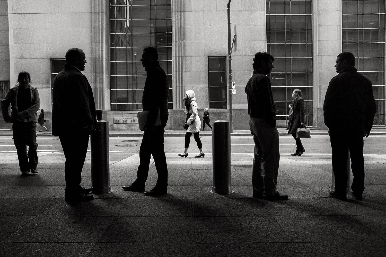 Black and white street photo of silhouetted people standing in line on a city sidewalk with bollards.