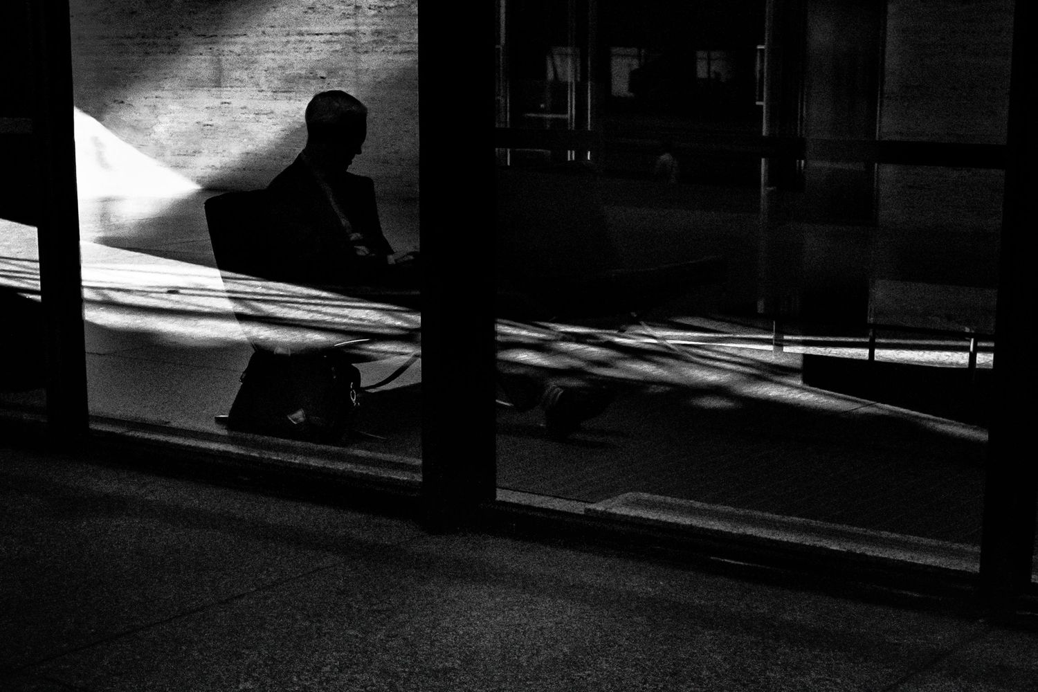 Black and white photo of a silhouetted person sitting alone in dramatic light and shadow near a glass building.