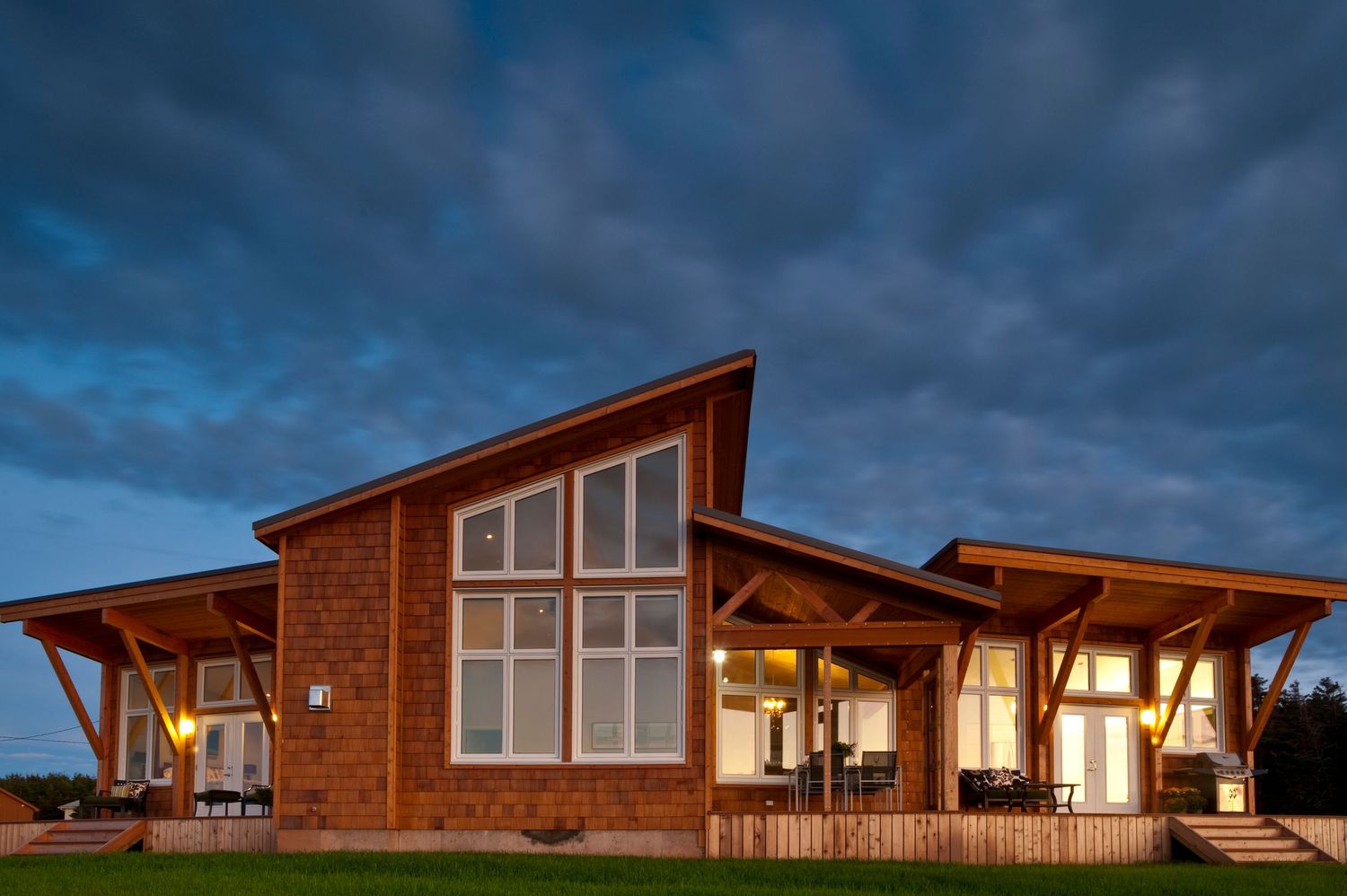 Modern timber frame house with large windows illuminated at dusk under dramatic cloudy sky.