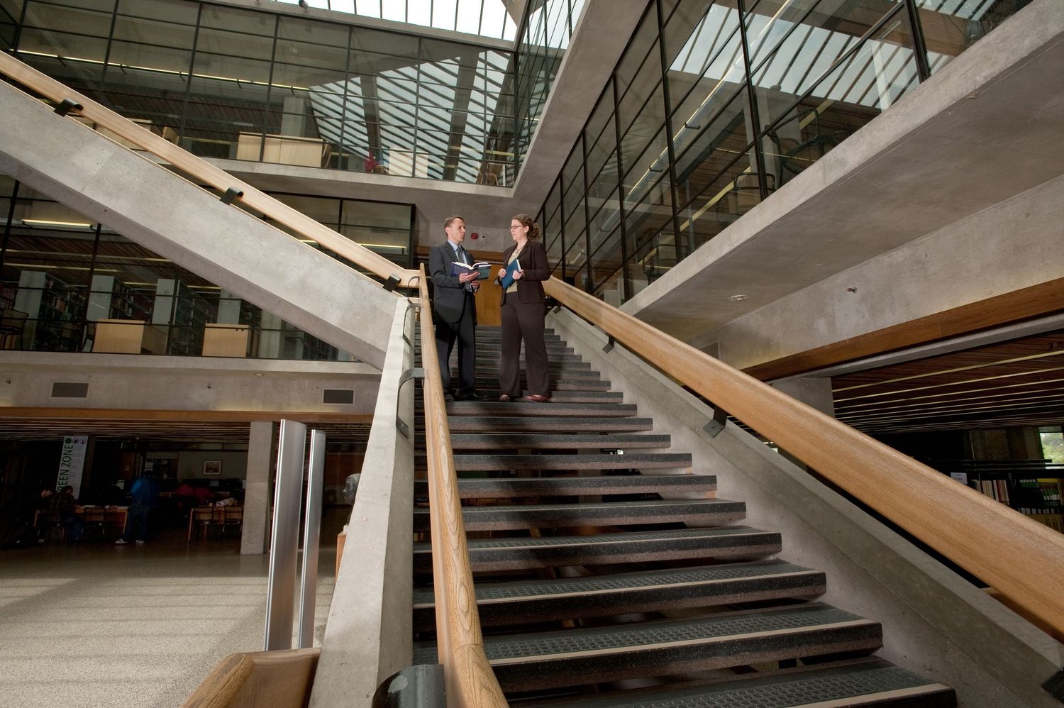 Two businessmen in suits descend a modern wooden staircase inside a glass-walled corporate building.