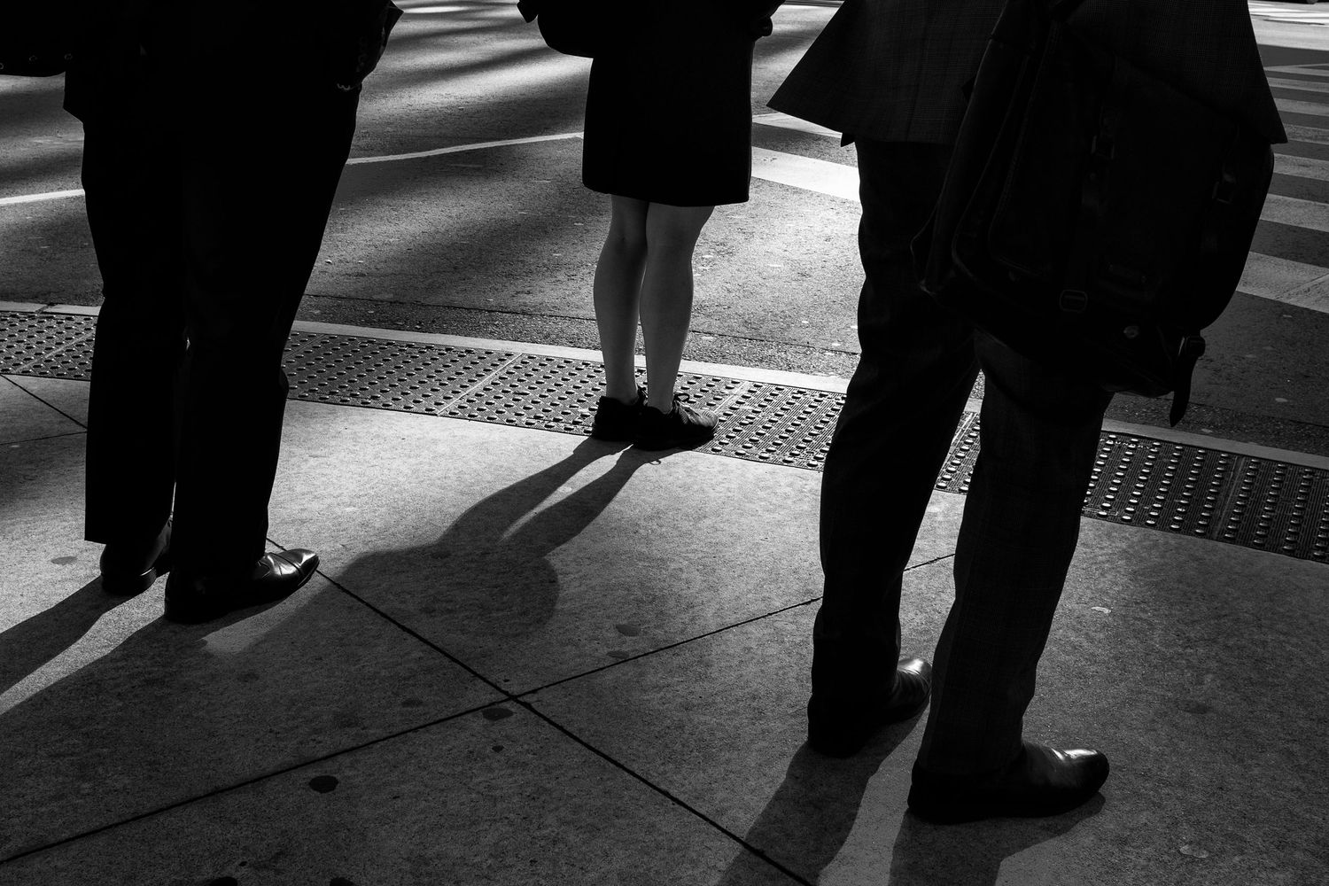 Black and white photo of people standing on a city sidewalk, casting dramatic shadows on the pavement.