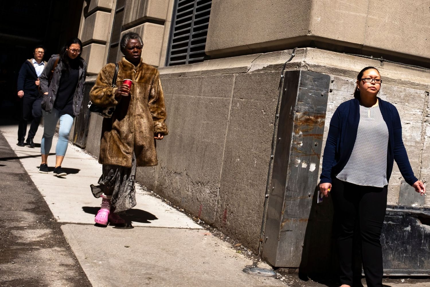 People walking on a city sidewalk, one person wearing a fur coat holding a red cup, another in a blue cardigan.
