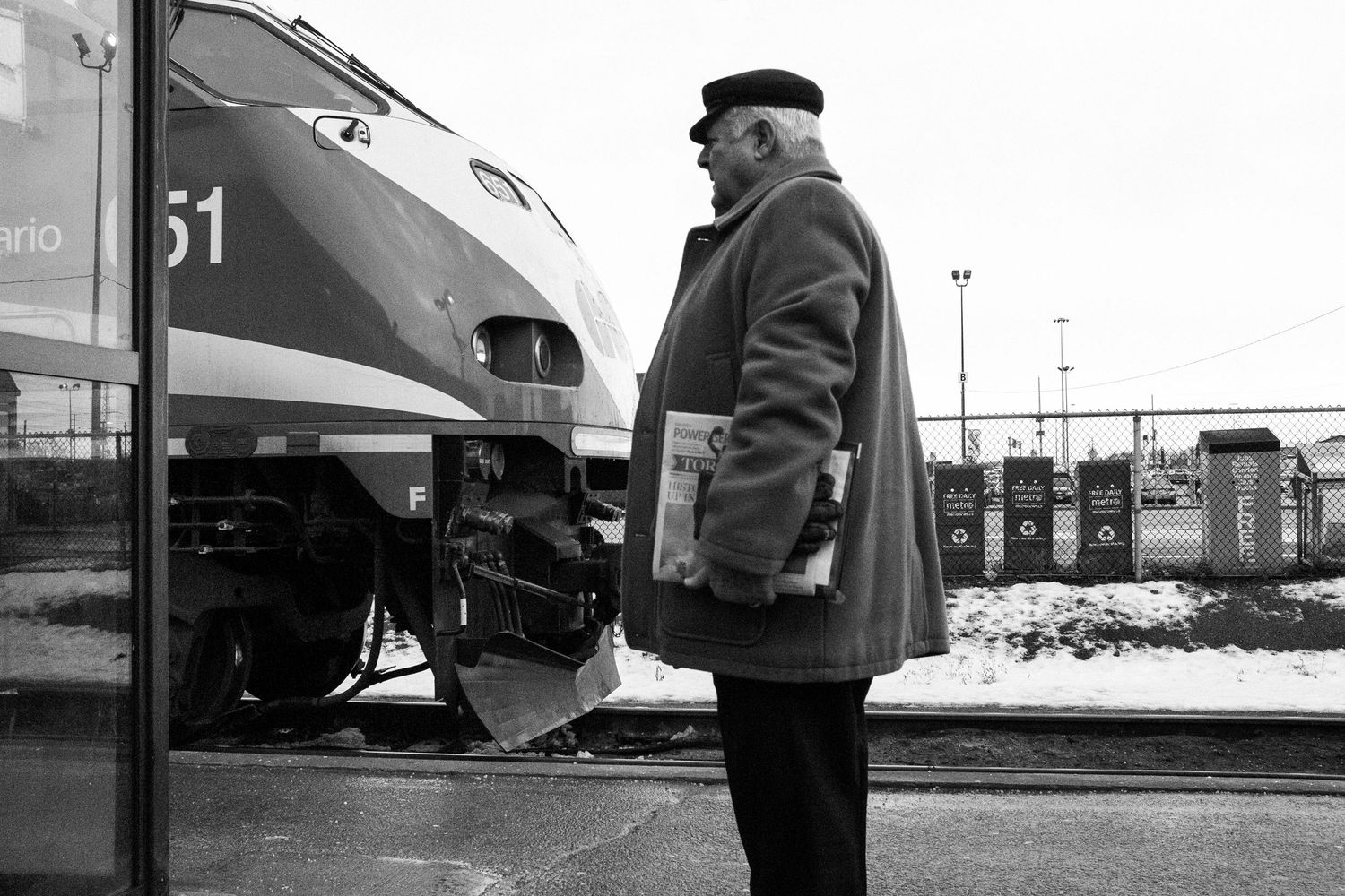 Elderly man in overcoat holding newspaper stands on snowy train platform beside modern commuter train, black and white photo.