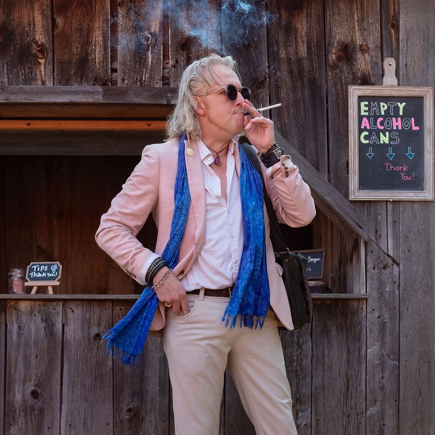 Stylish man in pink blazer and blue scarf smoking a cigarette outside a rustic wooden building.