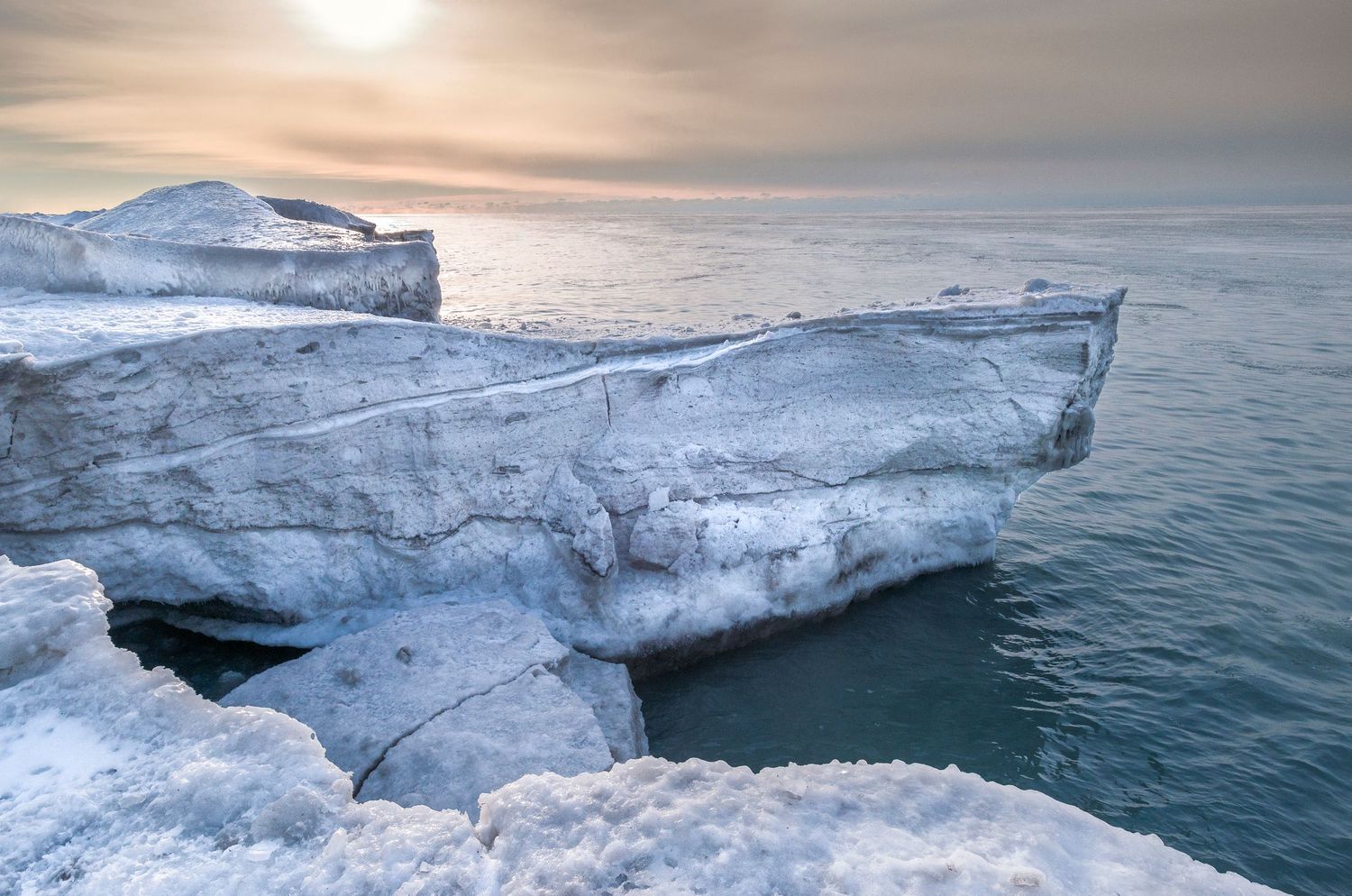 Icy cliffs overlooking a calm sea under a hazy winter sunset in a frozen Arctic landscape.