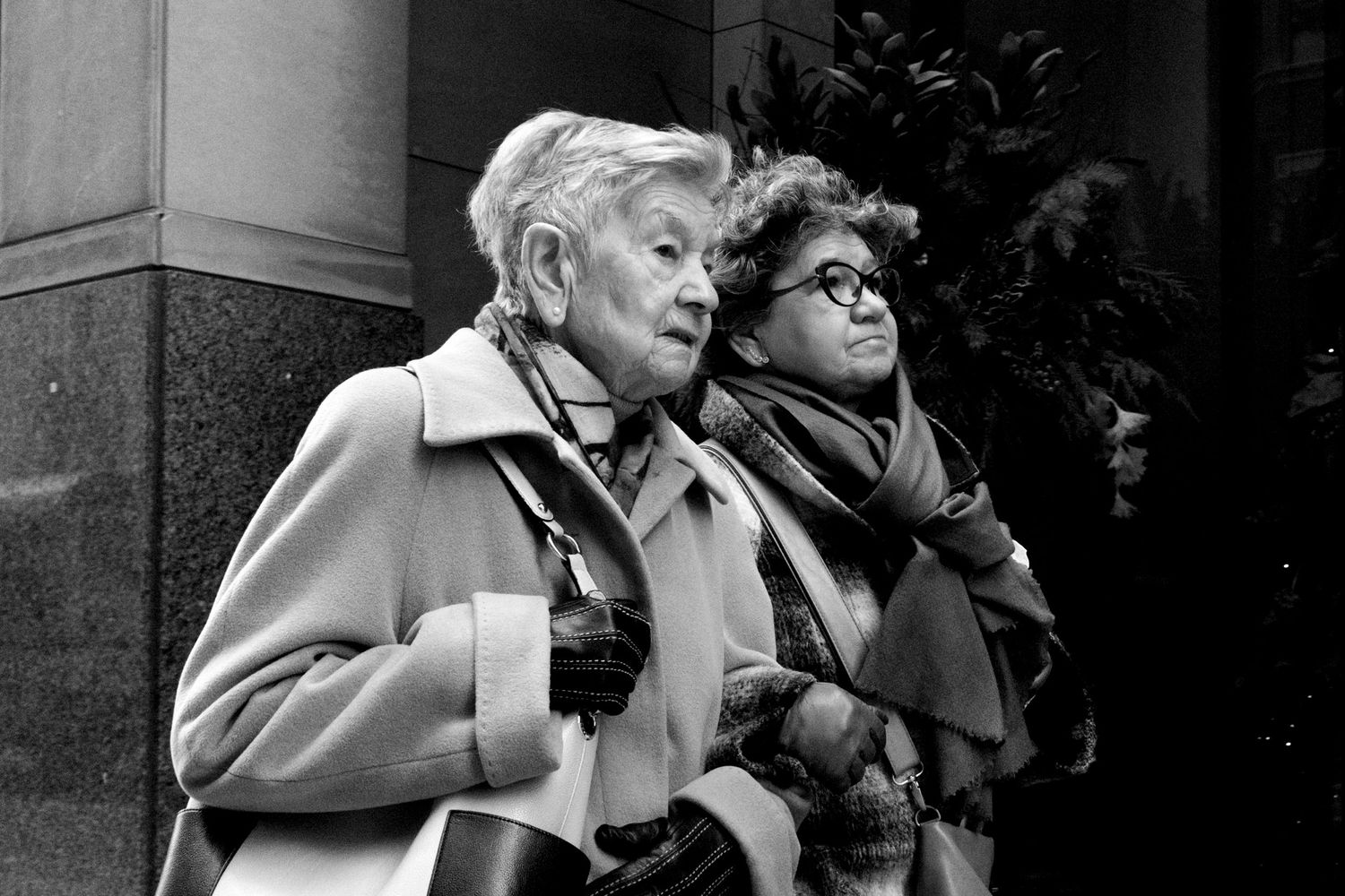 Black and white street photo of two elderly women in winter coats walking together in the city.