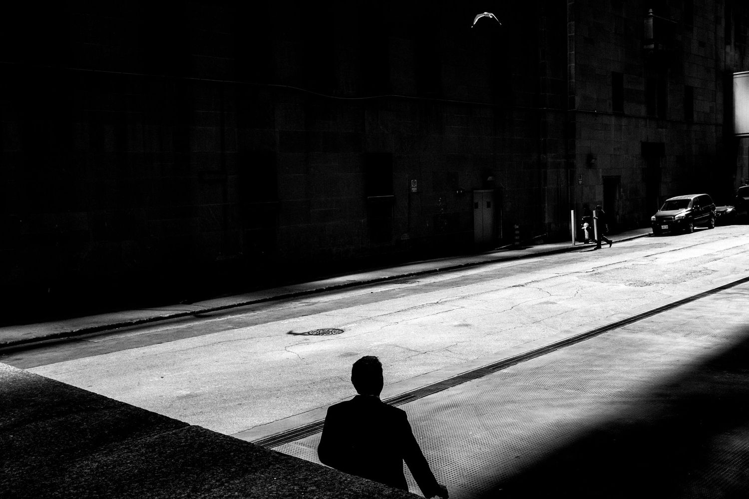 A solitary silhouette stands in a dramatic black and white urban street scene with stark light and shadow contrasts.