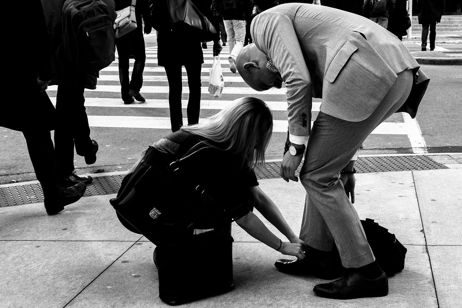 Black and white photo of two people on a city sidewalk, one bending down to help the other with luggage.