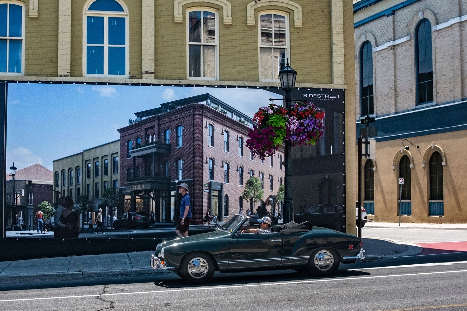 Vintage convertible car drives past a large outdoor mural on a sunny urban street.