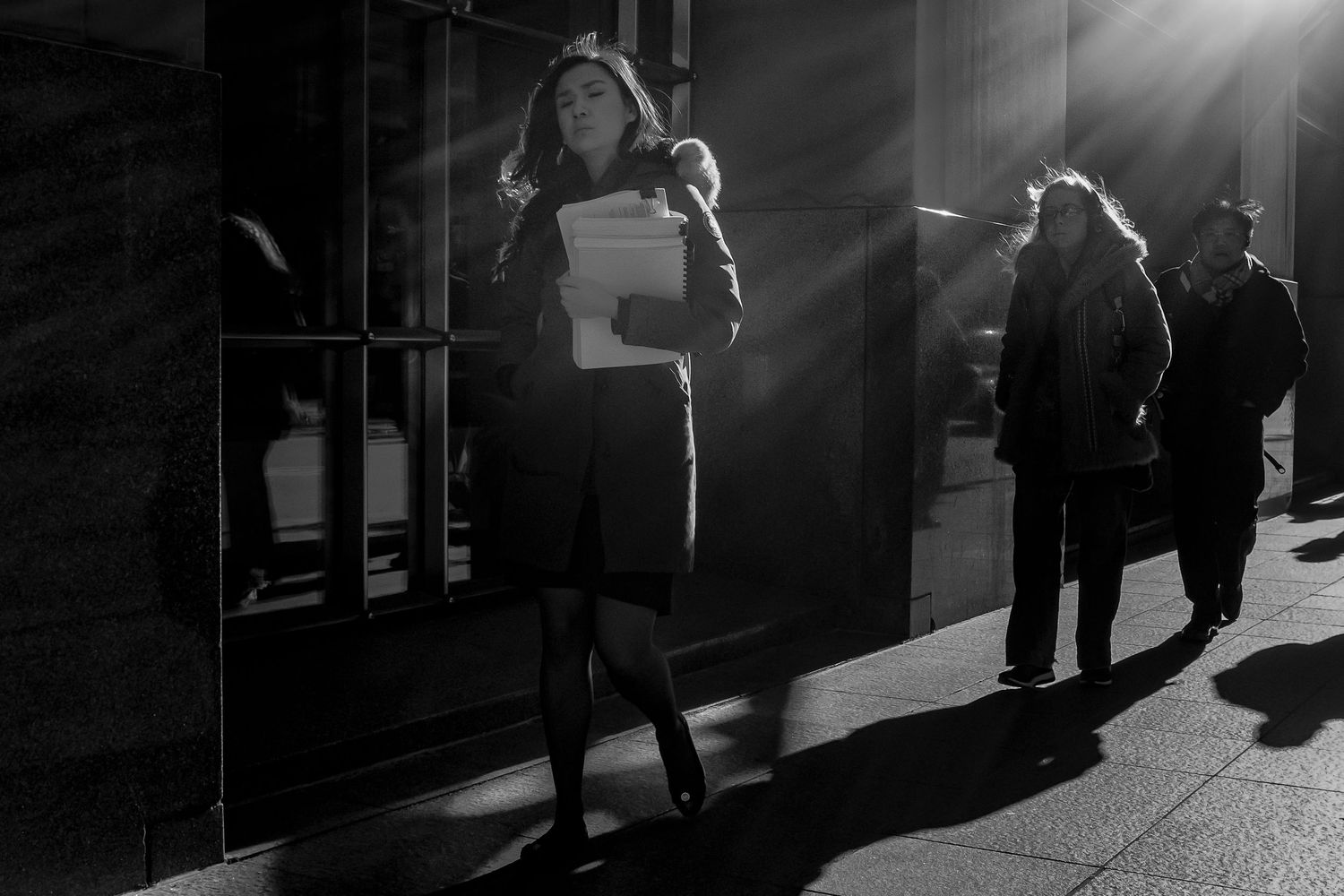 Black and white street photo of a woman holding papers in dramatic sunlight, with two people walking behind her.