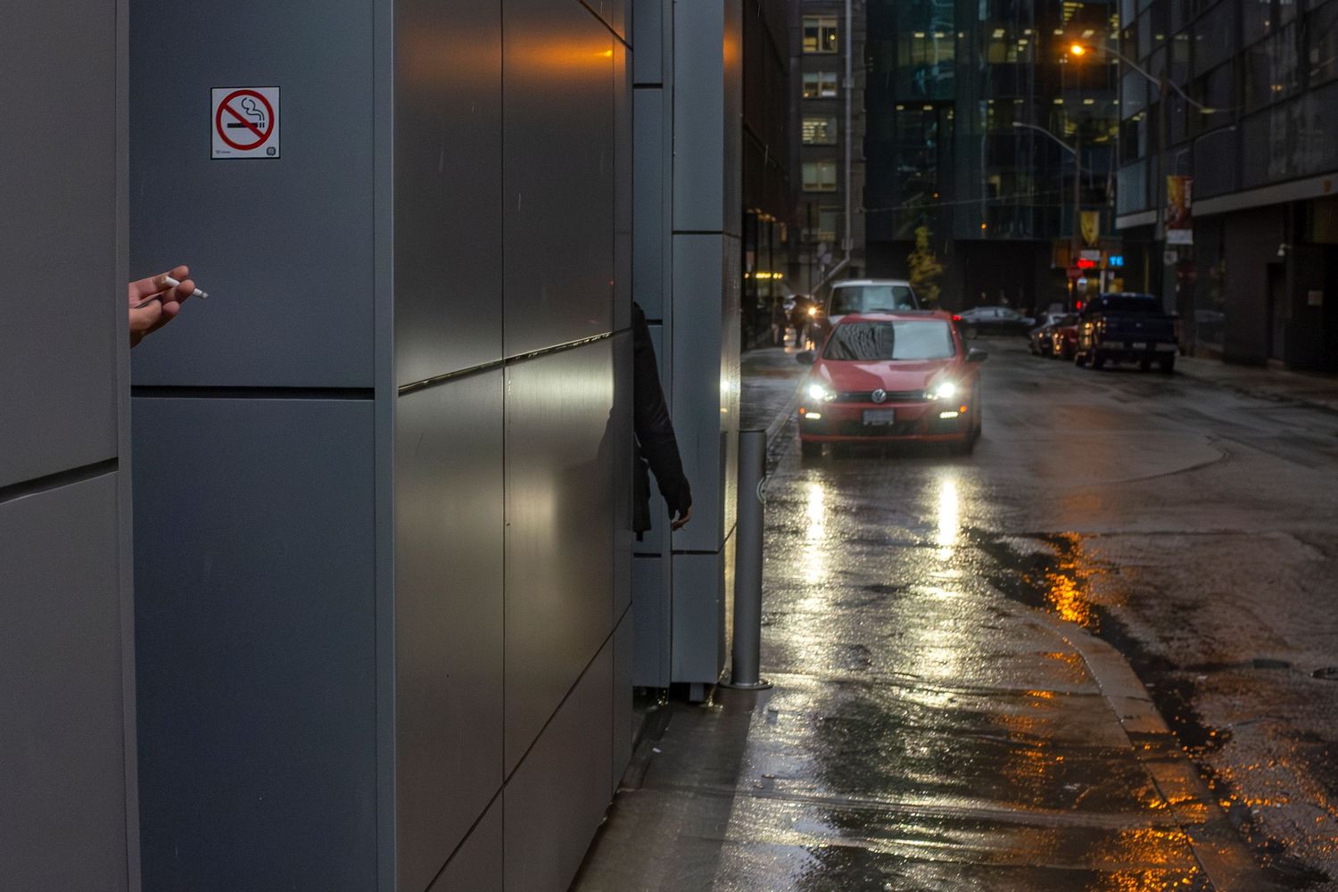 Rainy city street at dusk with a red car, wet reflective pavement, and a hand holding a cigarette near a no-smoking sign.