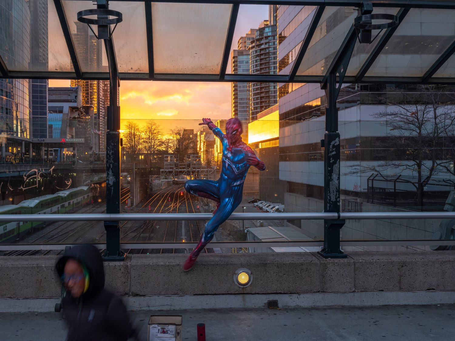 Spider-Man costume performer poses dramatically at a city bus stop at sunset with urban skyline in background.