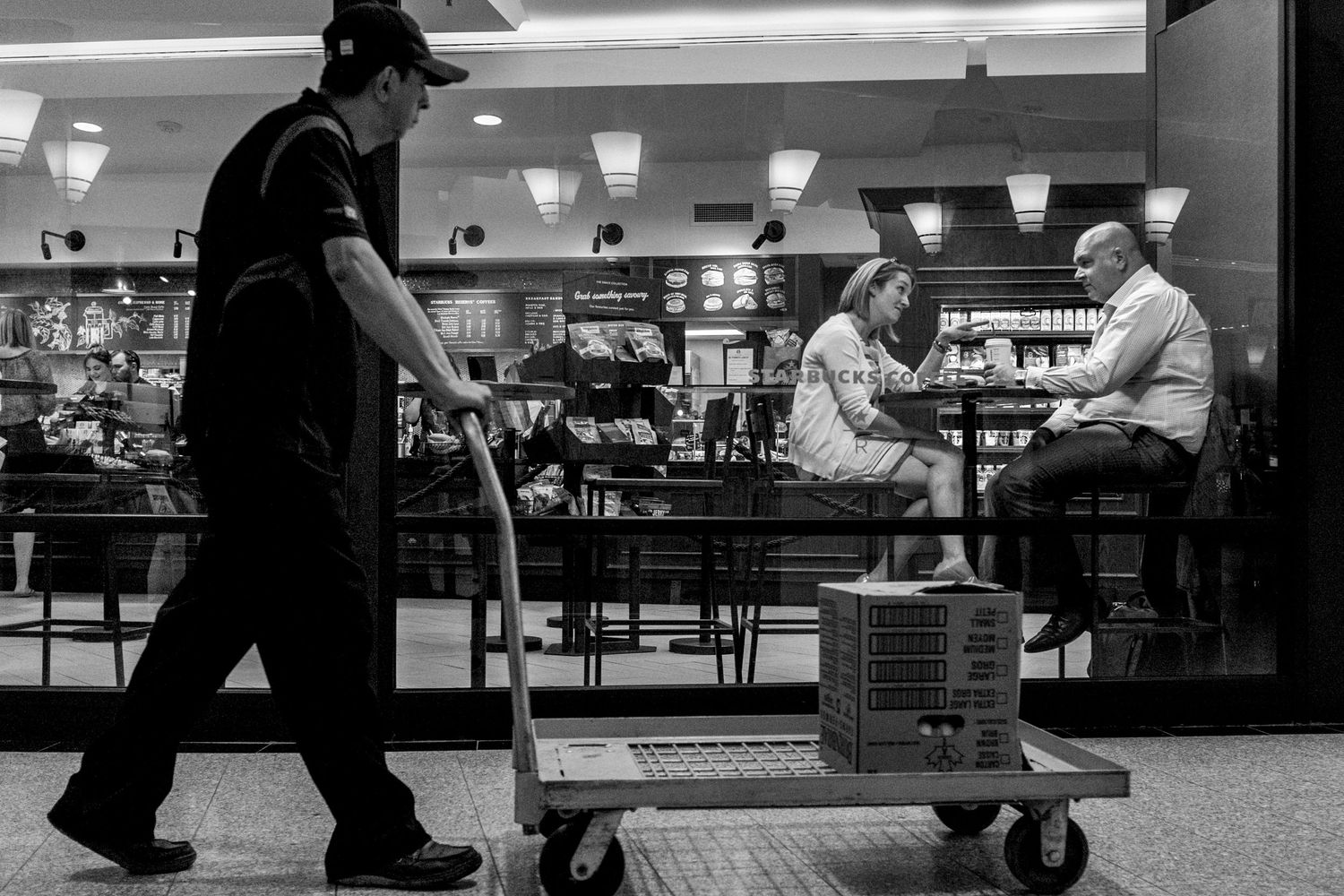Black and white photo of a worker with a cart passing diners at a restaurant in a busy setting.