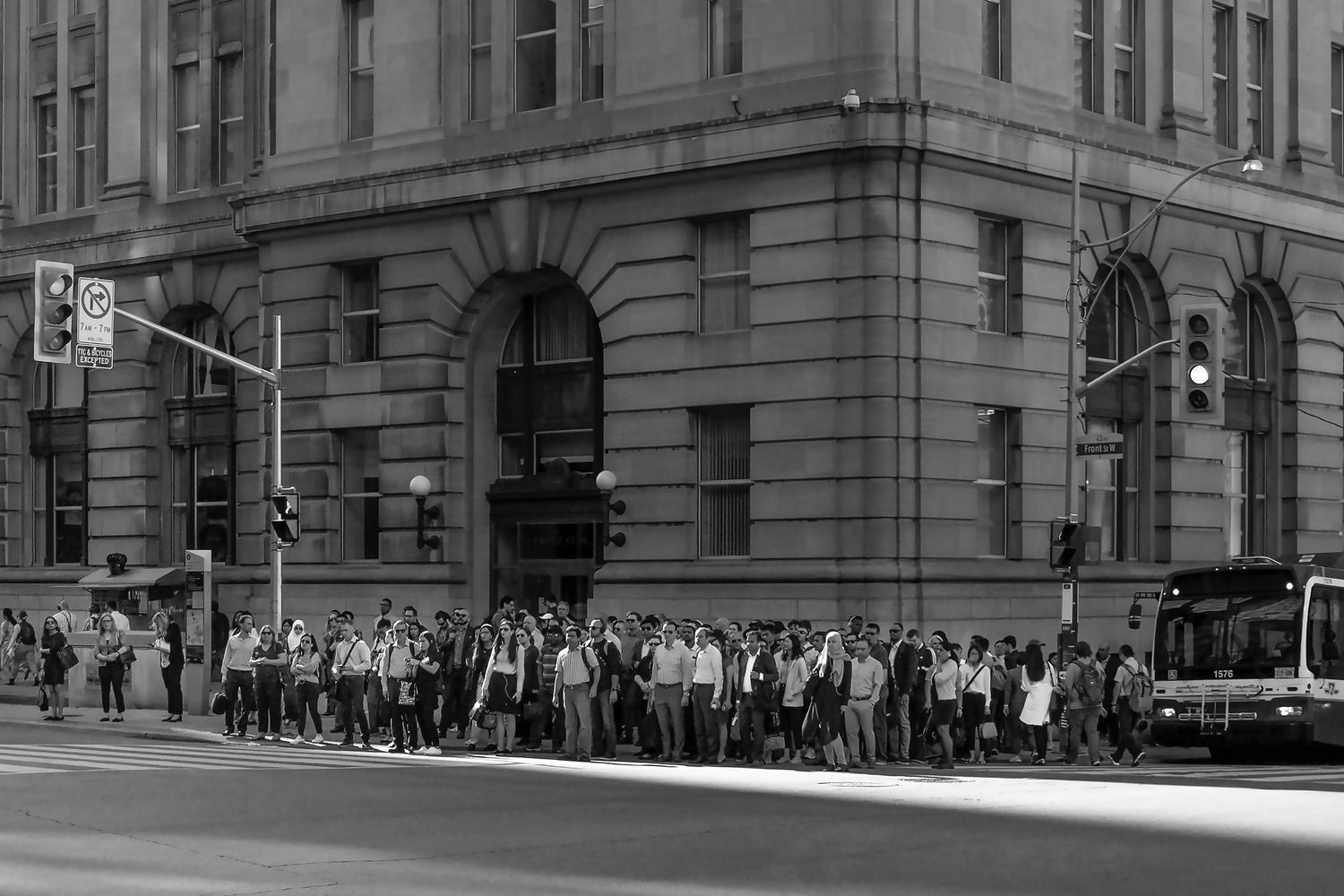 Black and white photo of a large crowd of people waiting at a bus stop outside a grand stone building in a city.