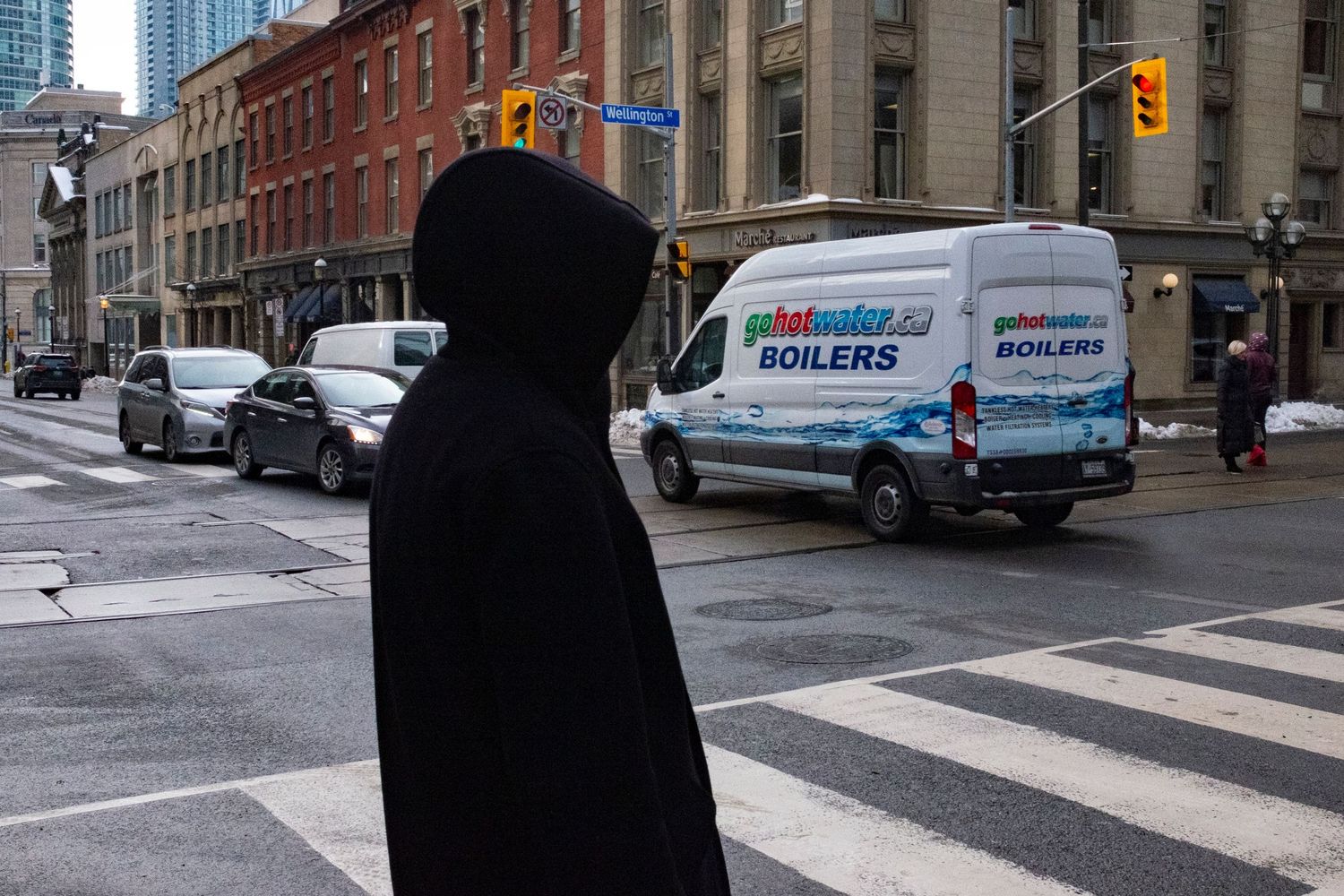 Person in black hoodie standing at urban intersection with a Polvwater Boilers van driving through a city street.