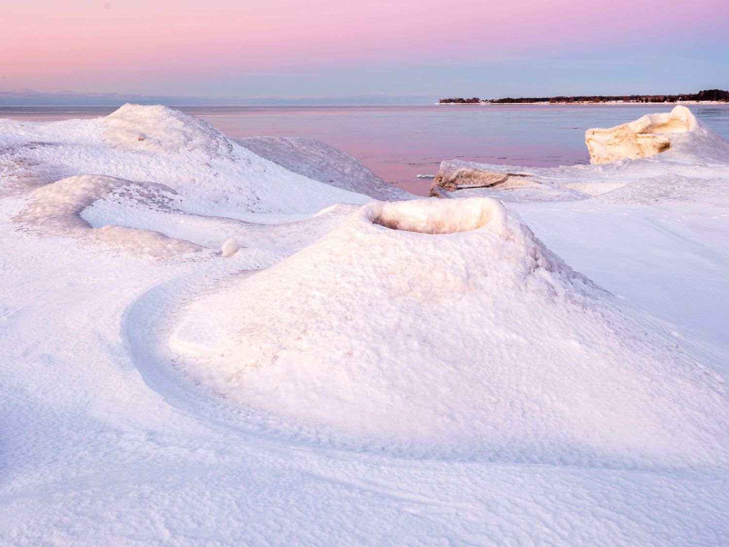 Snow-covered mounds along a frozen shoreline glow under a soft pink and purple twilight sky over calm waters.