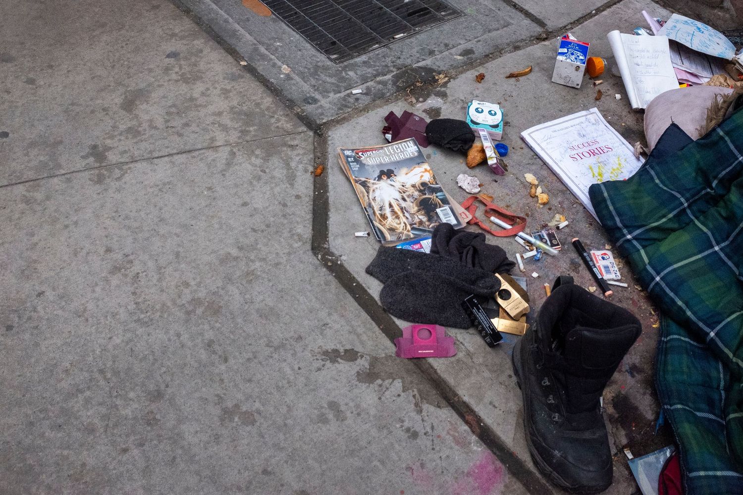 Scattered belongings on a sidewalk including books, shoes, and miscellaneous items next to a plaid blanket.