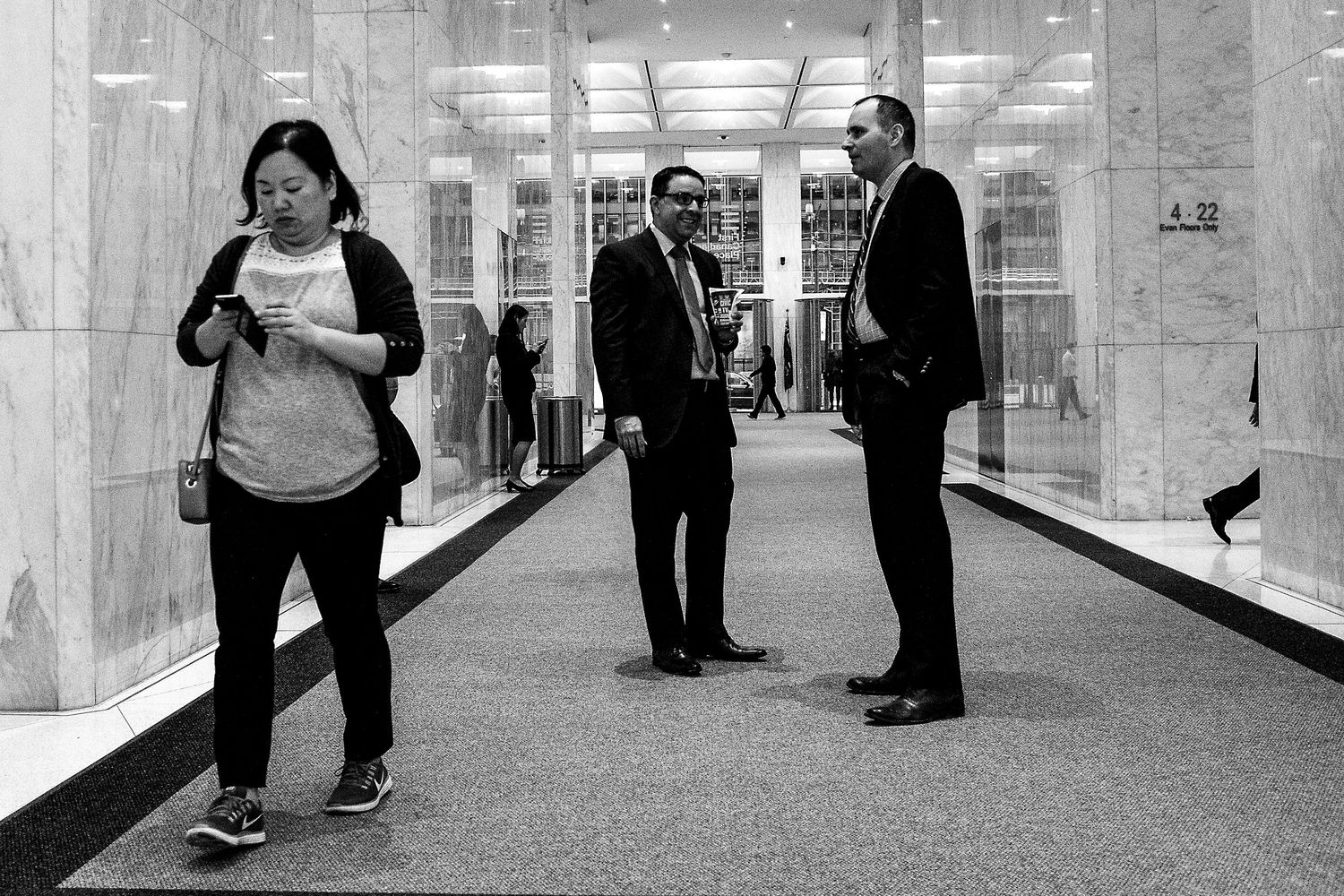Black and white street photo of three people in a modern building lobby, each in their own world.