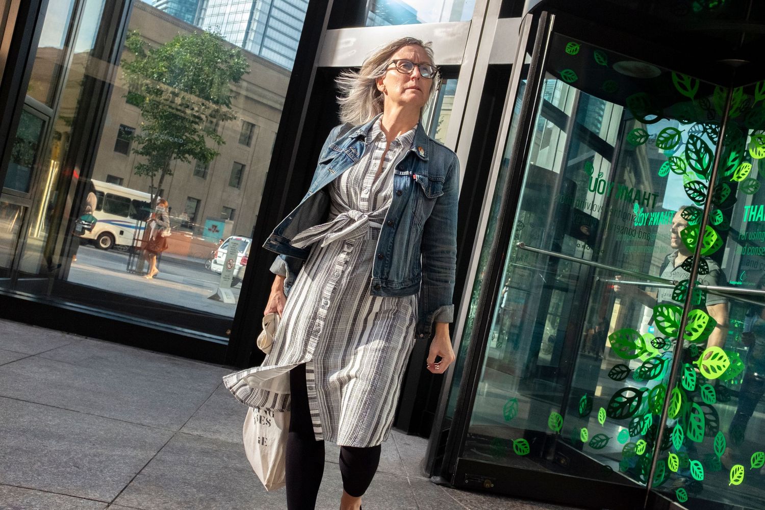 Stylish woman with silver hair wearing a denim jacket and striped dress walking outside a modern building.