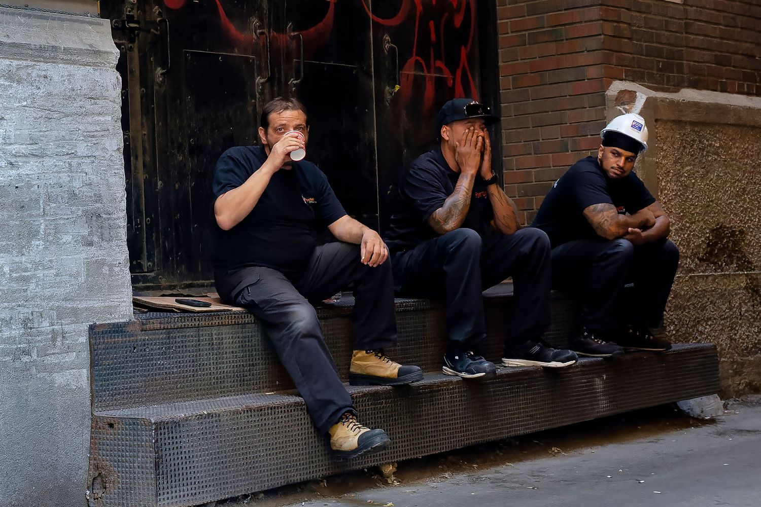 Three men sitting on urban steps, one drinking coffee, another with head in hands, in a city alleyway.