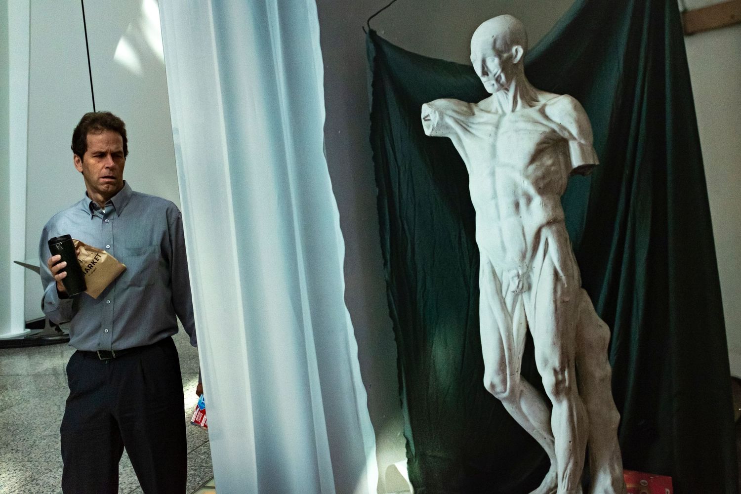 A man holding flowers stands beside a detailed anatomical human figure sculpture draped in green fabric.