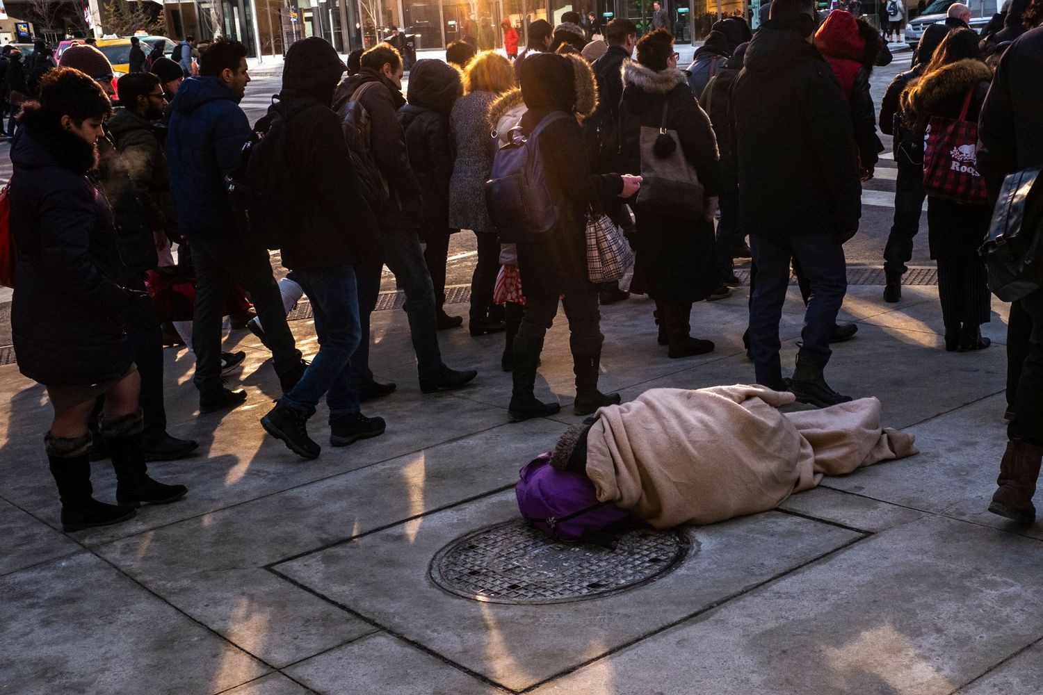 A person wrapped in a blanket lies on a city sidewalk surrounded by pedestrians at sunset.