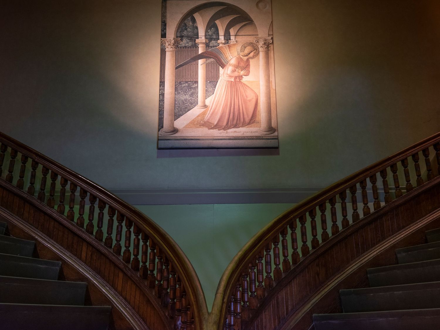 Elegant split staircase with wooden railings and a Renaissance-style angel fresco illuminated on a green wall.