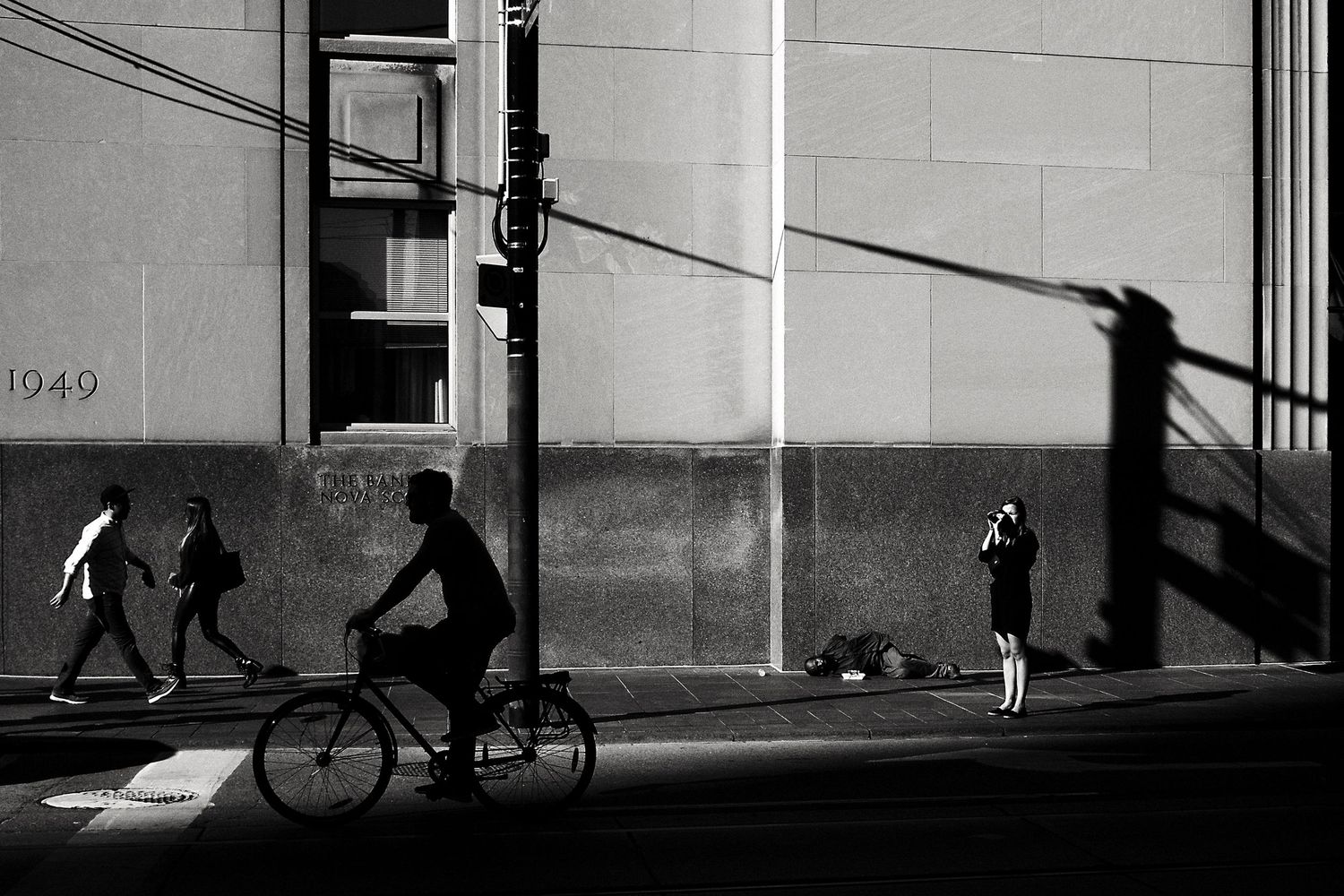 Black and white street photo of cyclist, pedestrians, and a photographer near a building marked 1949 with dramatic shadows.
